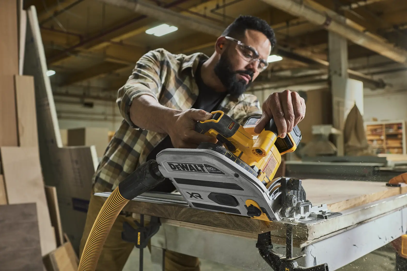 Une personne utilise une scie circulaire sans fil DEWALT pour couper du bois sur un établi dans un atelier. La scie est connectée à un tuyau d'extraction de poussière. Le visage de la personne est flouté pour préserver sa confidentialité.