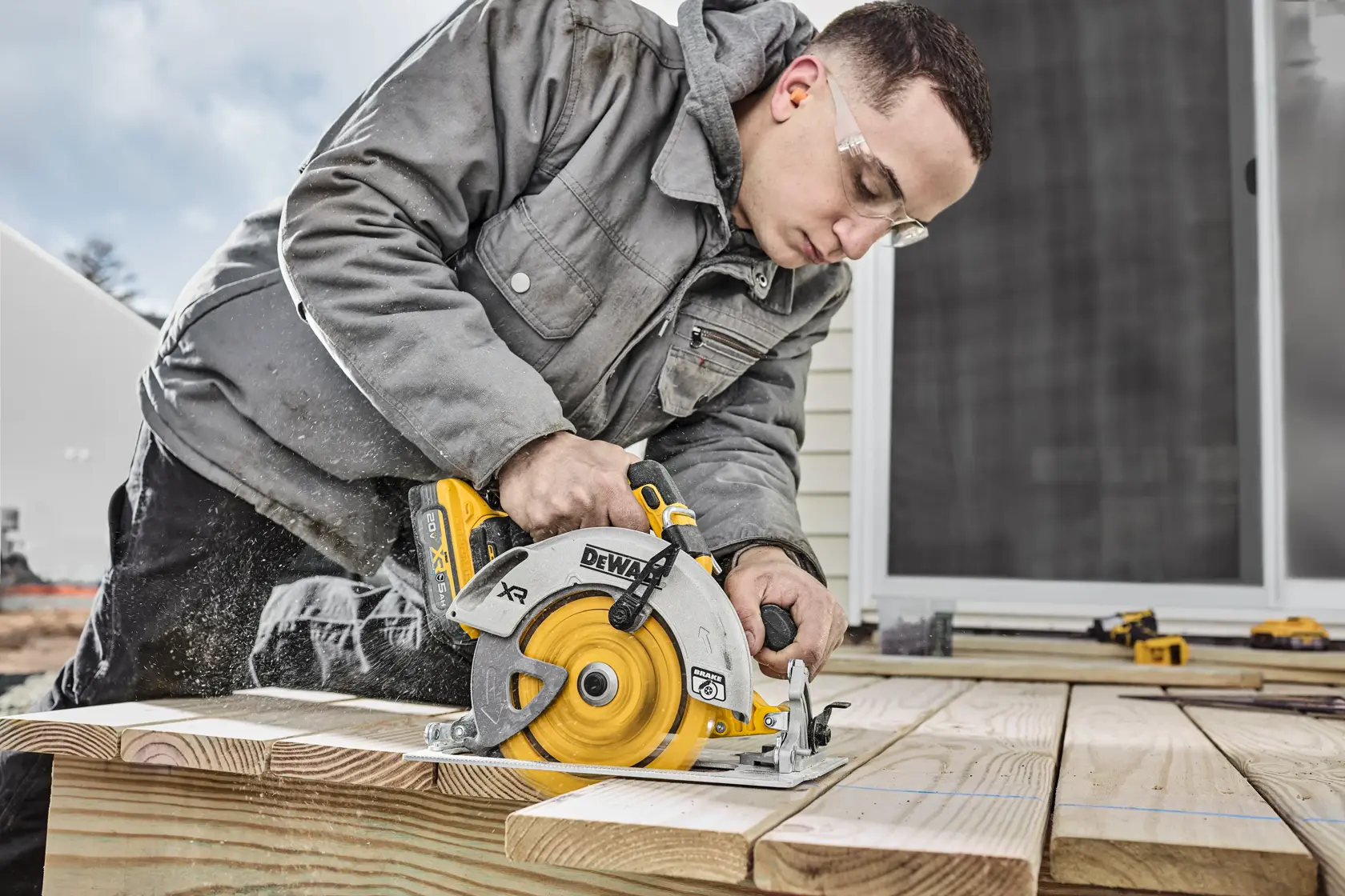 A person using a DEWALT cordless circular saw to cut wooden planks outdoors. The worker is wearing a gray jacket and the saw is producing sawdust. Other tools are visible on the wooden surface.