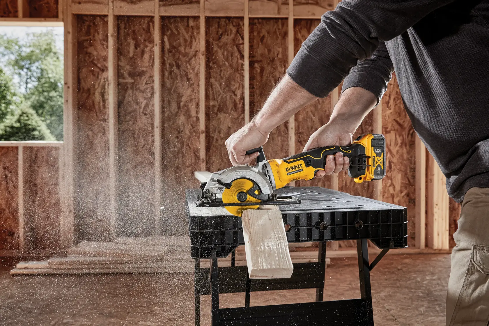 A person using a DEWALT cordless circular saw to cut a piece of wood on a workbench inside a partially constructed building.