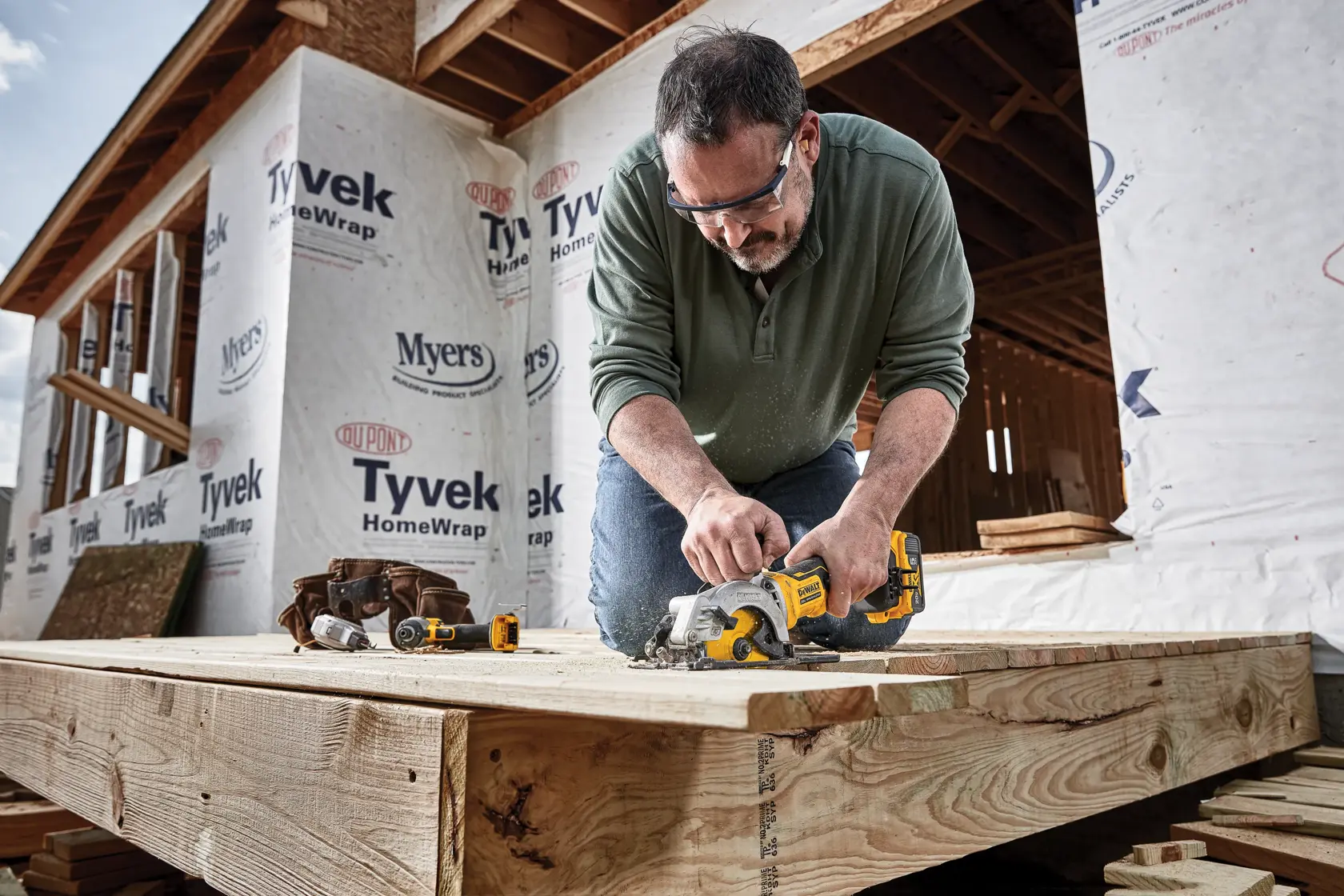 A person uses a DEWALT circular saw to cut a wooden plank at a construction site. The individual is kneeling on the wood deck in front of a building under construction, with Tyvek HomeWrap visible on the walls and additional DEWALT tools and a tool belt nearby.