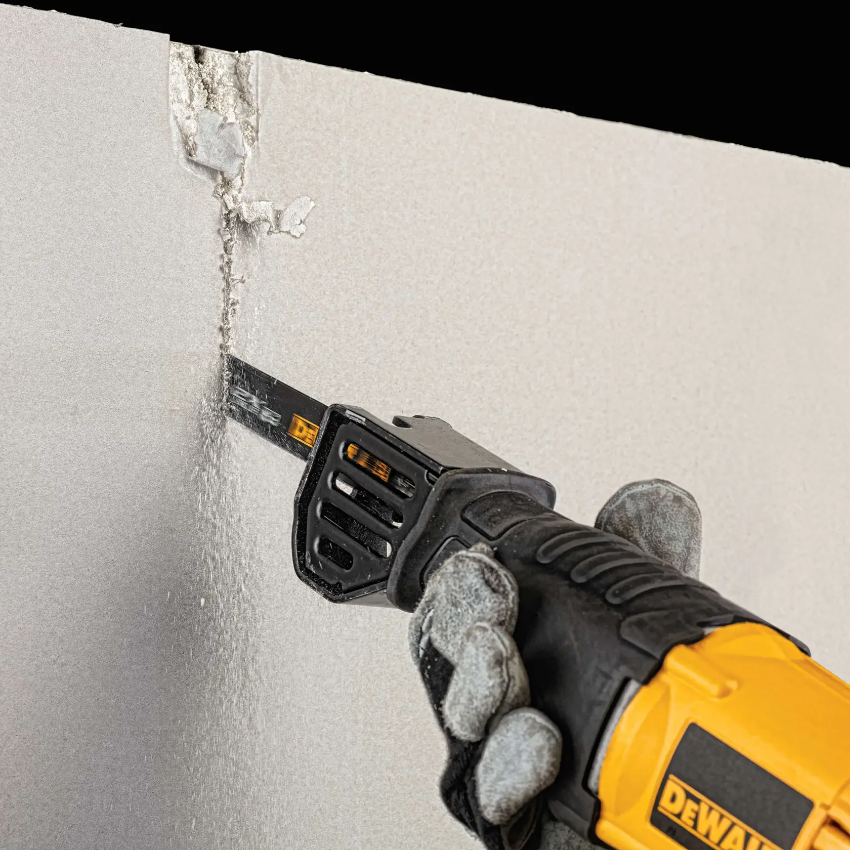 A gloved hand is using a DEWALT reciprocating saw to cut through a piece of drywall, with debris and dust visible along the cut.