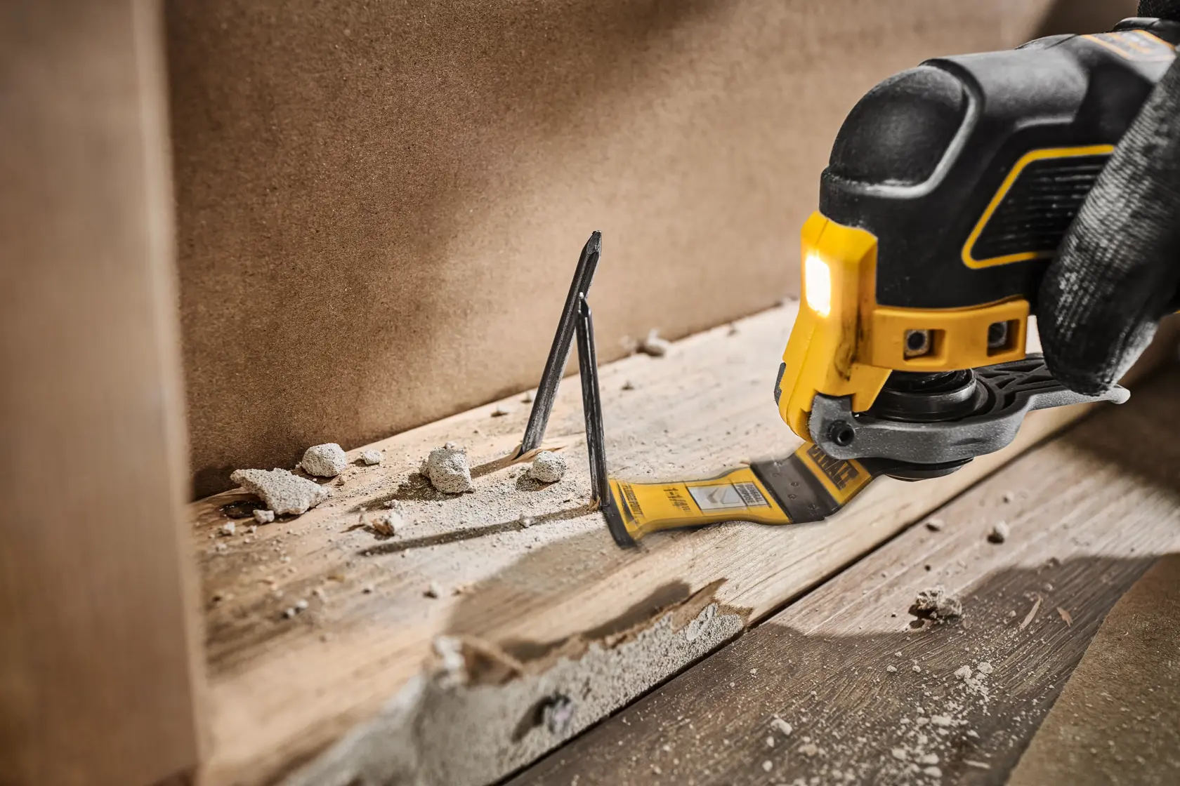 A DEWALT oscillating multi-tool is being used to cut through nails embedded in a wooden board, with dust and debris scattered around the work area.