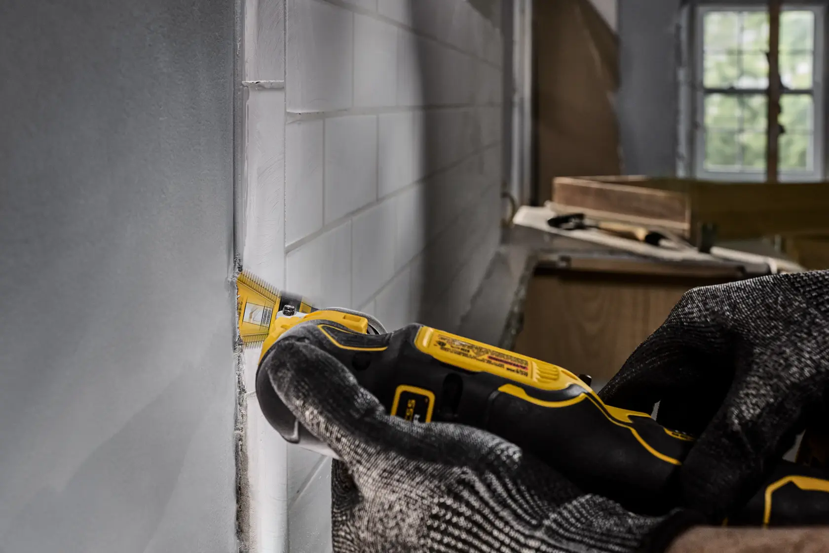 A person wearing protective gloves uses a DEWALT oscillating tool to cut or trim around white wall tiles during renovation or construction work.