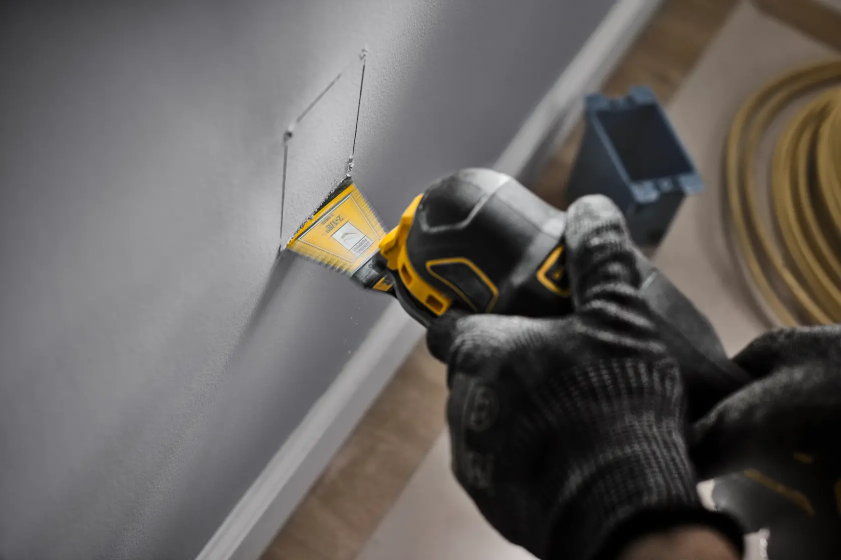 A person wearing gloves uses a DEWALT oscillating multi-tool with a yellow blade to cut a rectangular hole in a gray wall. Nearby, there are electrical components and coiled cables on the floor.