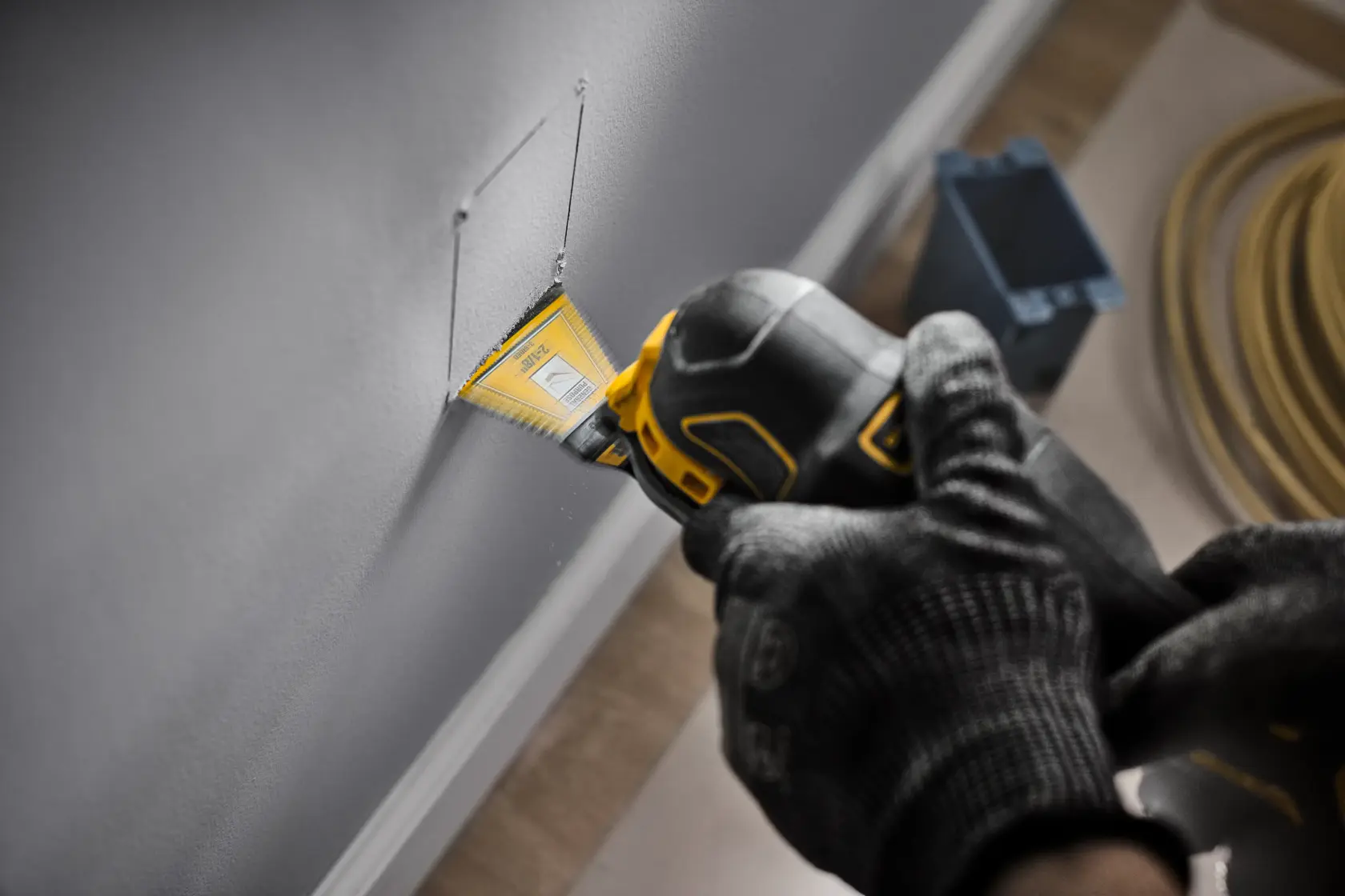 A person using a DEWALT oscillating multi-tool with a yellow blade to cut a square in drywall. The user is wearing black gloves and cutting a hole in the wall, likely for electrical installation. The tool and blade are clearly visible.