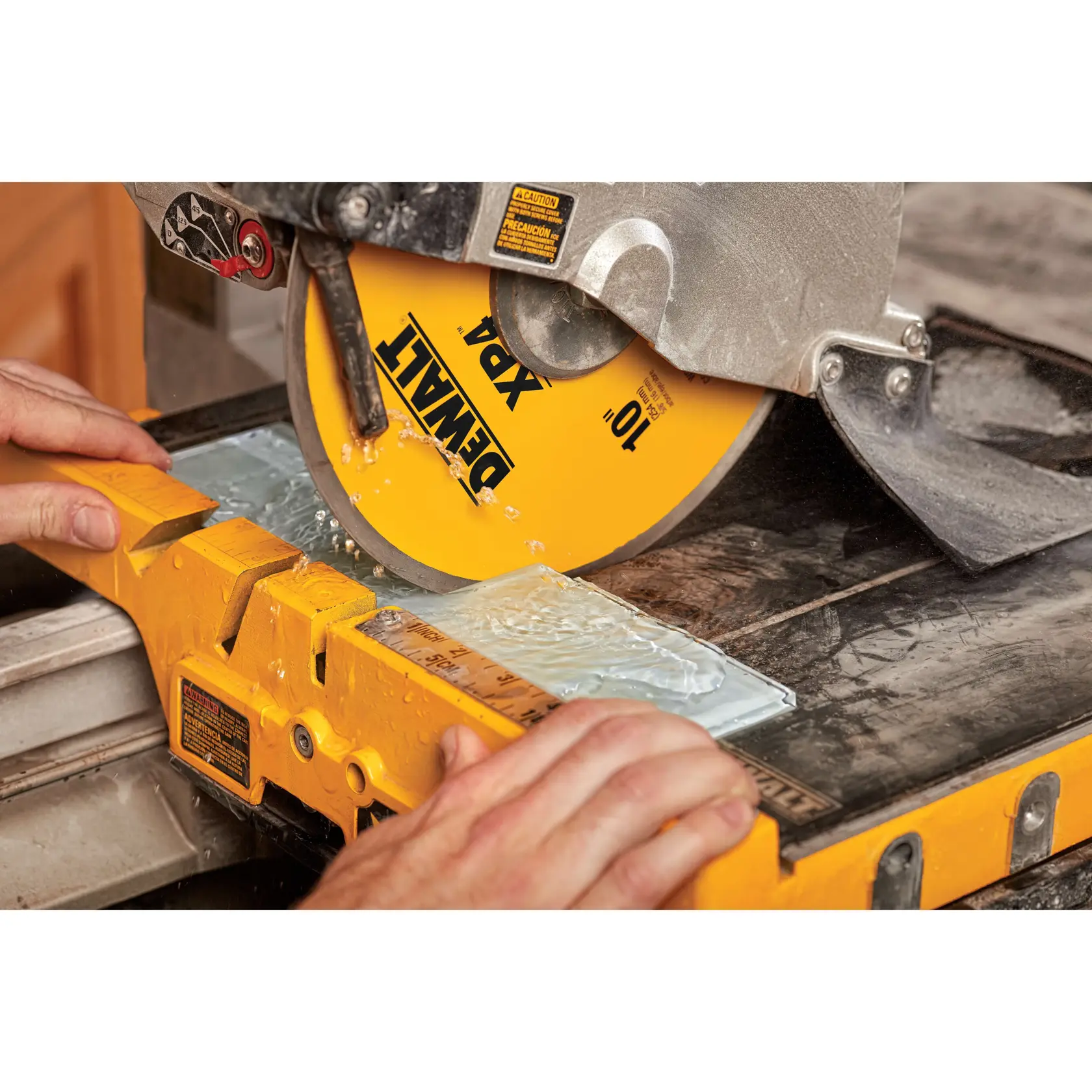 Close-up image of a DEWALT XP4 wet tile saw blade cutting a glass tile, with water flowing over the tile and hands holding the tile in place on the saw's yellow base.