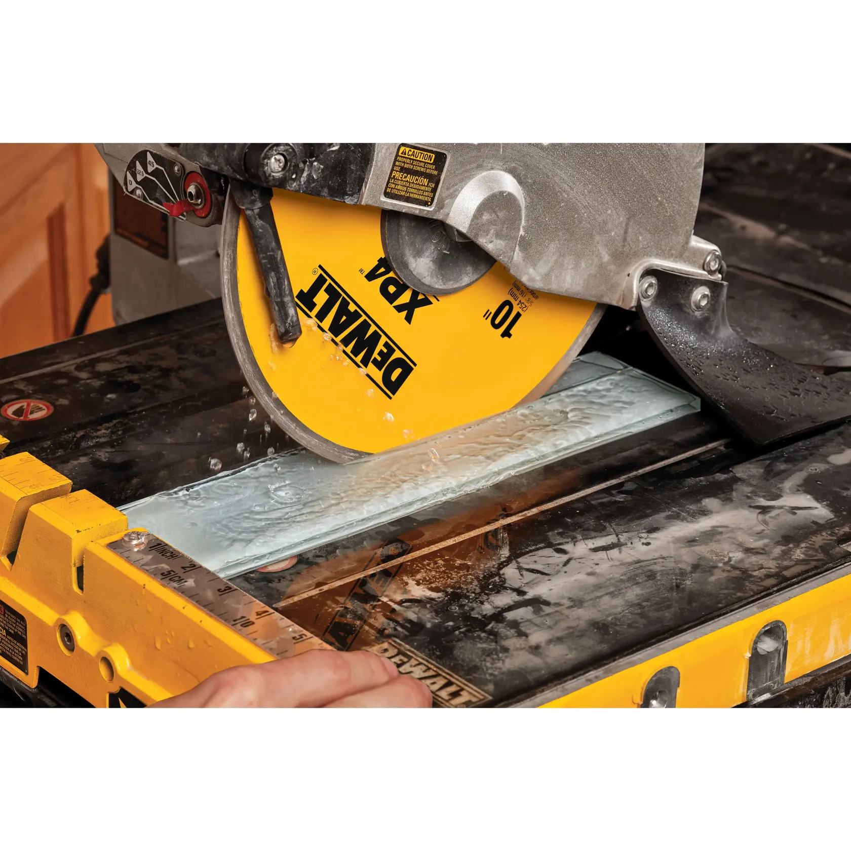 A DEWALT branded yellow circular saw blade cutting a piece of tile on a wet saw table, with water flowing to cool the blade.