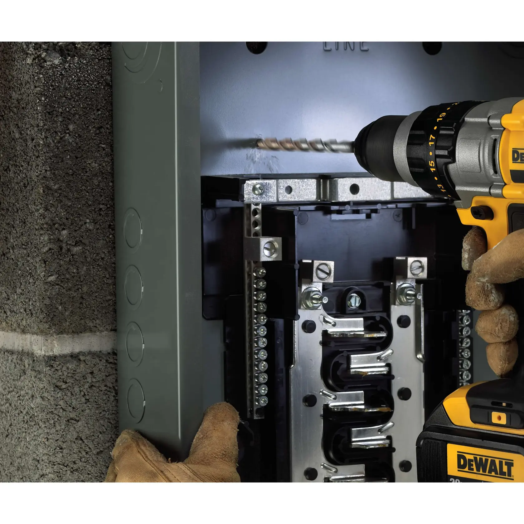 A person wearing brown gloves is using a DEWALT cordless drill to make a hole in a metal panel inside an electrical breaker box mounted on a concrete wall.