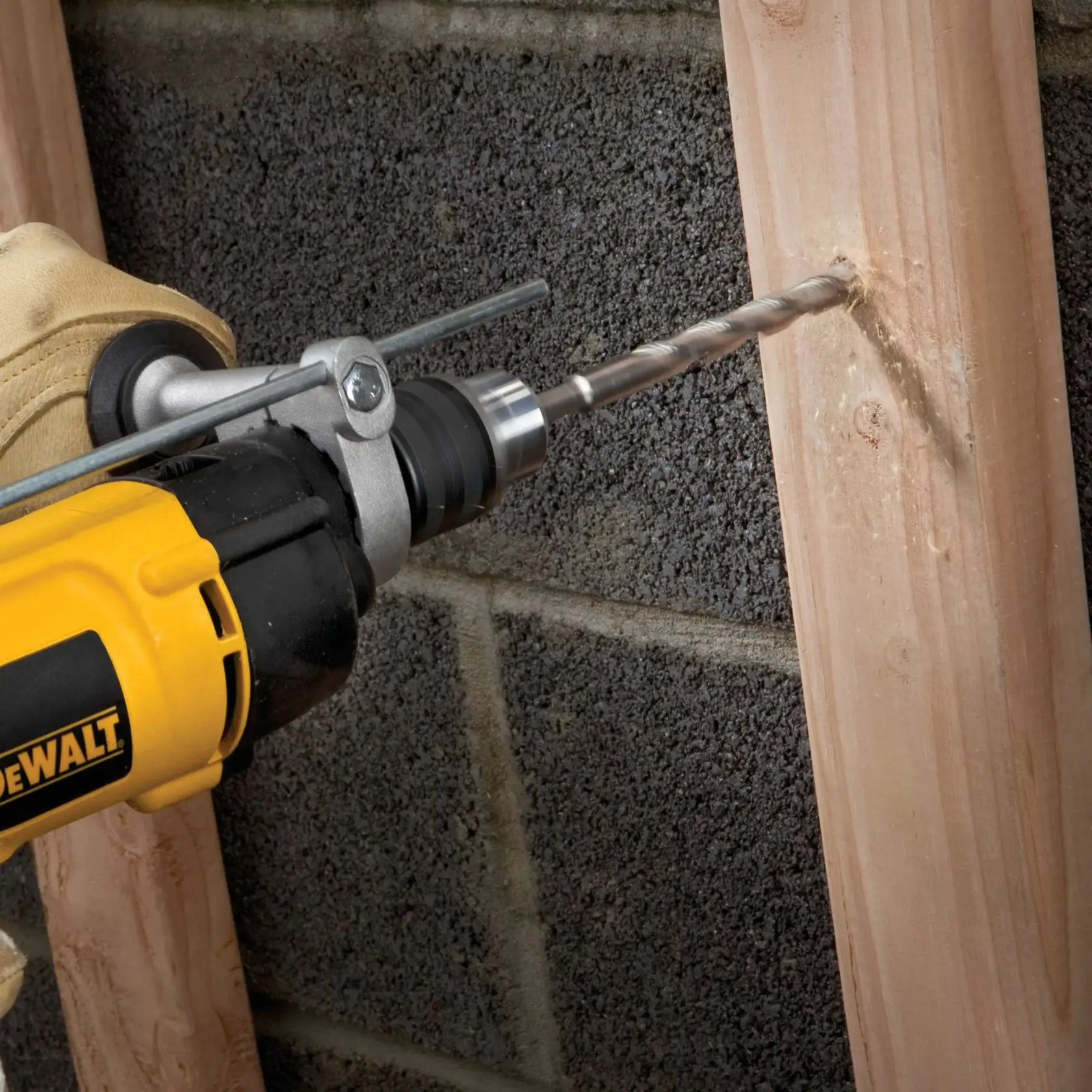 Close-up of a yellow DEWALT drill being used to drill into a wooden beam, held by a gloved hand, with a concrete block wall in the background.