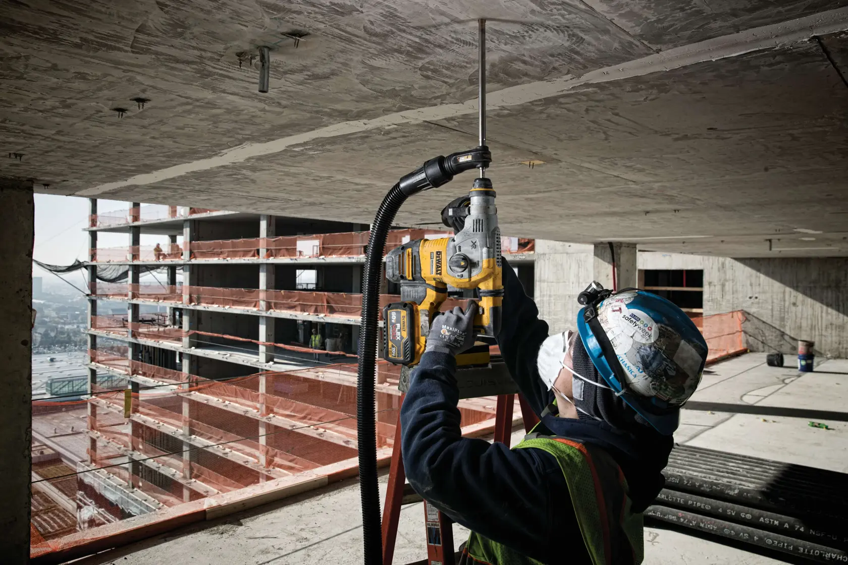 A construction worker wearing protective gear uses a DEWALT power drill to bore into a concrete ceiling at a building site, with a partially constructed structure visible in the background.