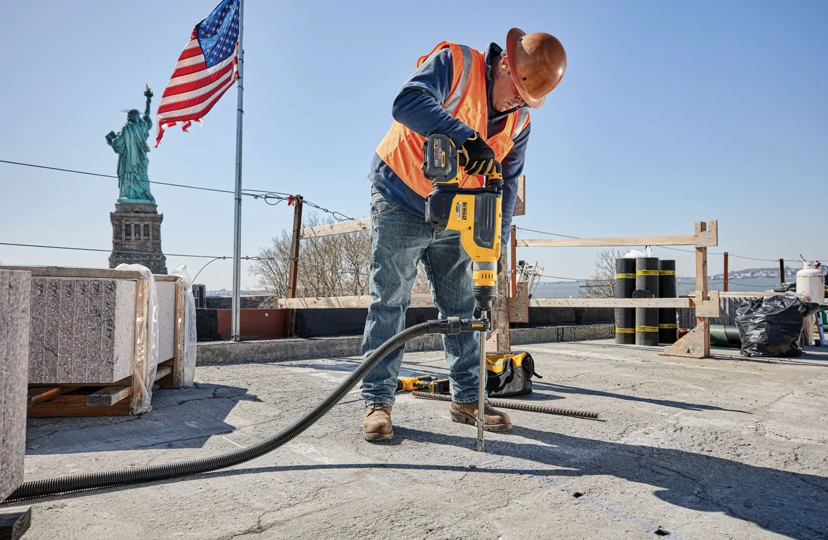 A construction worker wearing a hard hat and orange safety vest operates a yellow DEWALT power tool to drill into concrete on a job site. The Statue of Liberty and an American flag are visible in the background.