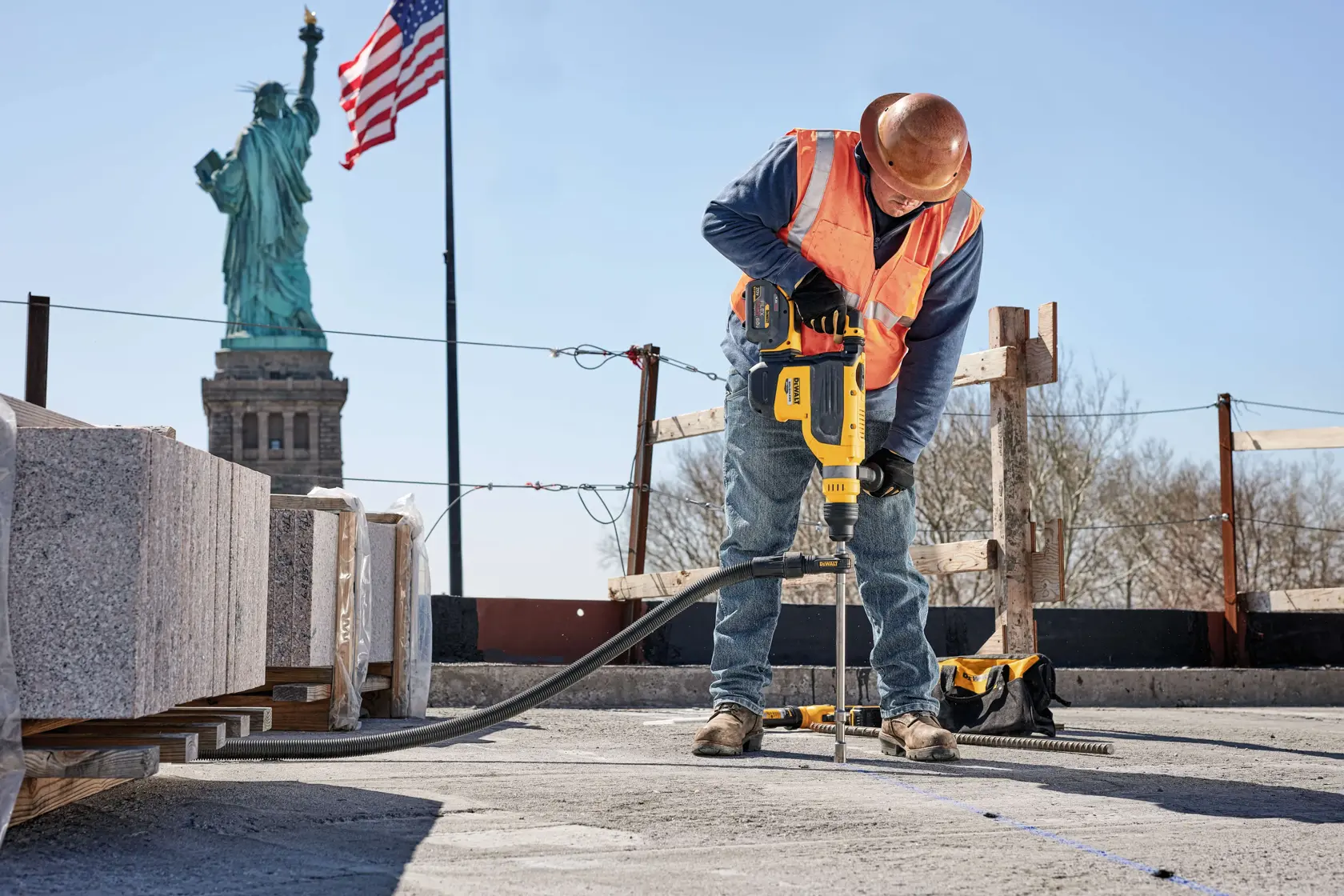 A construction worker uses a yellow DEWALT power tool to drill into concrete at a work site. The Statue of Liberty and an American flag are visible in the background.
