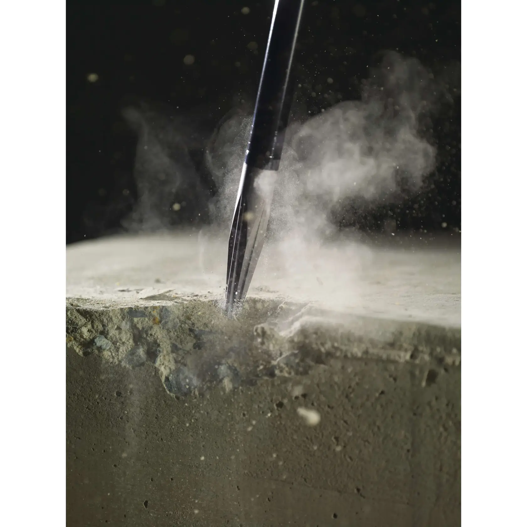 A close-up of a pointed chisel tool breaking into a concrete block, with dust and debris visibly flying around.
