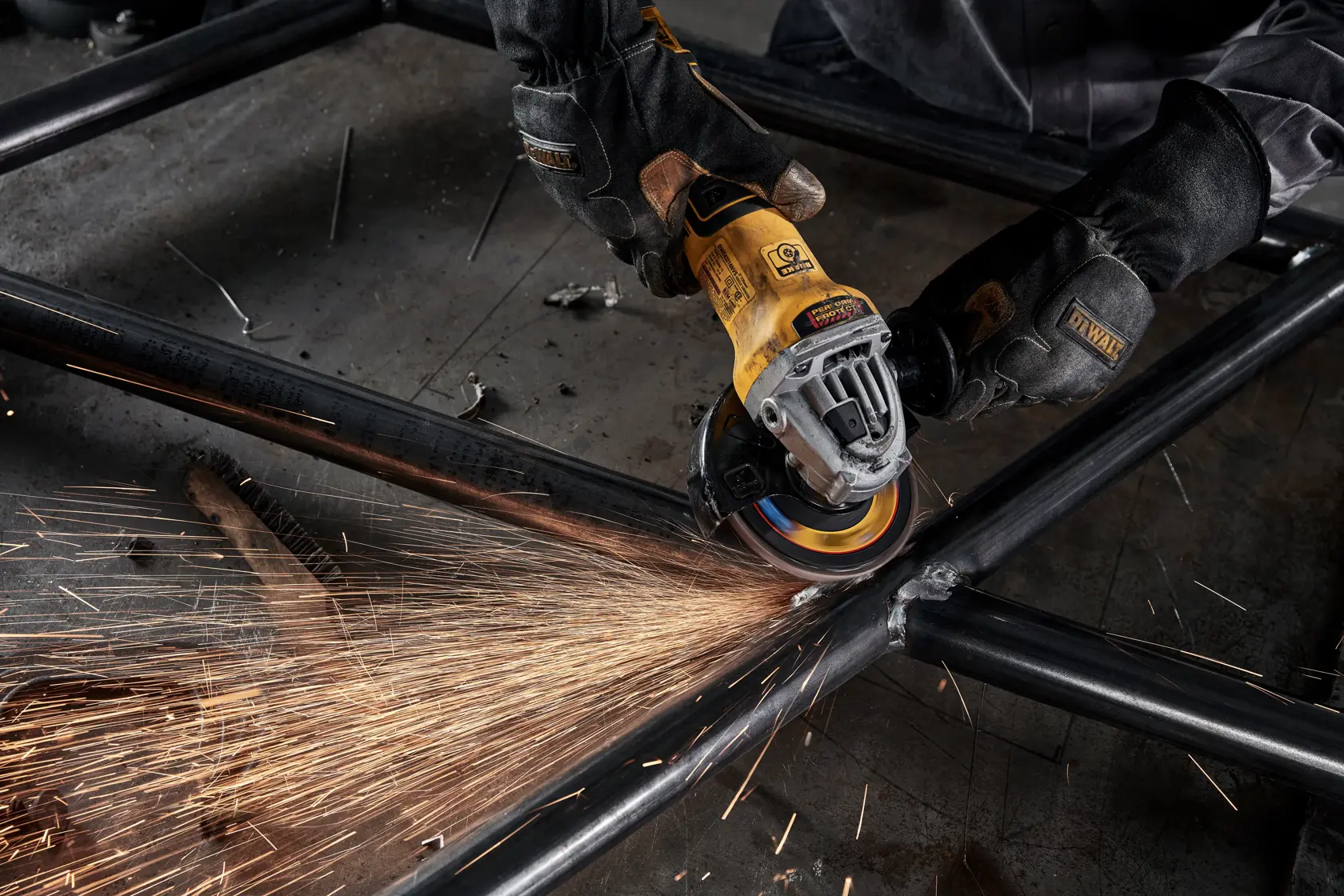 A person wearing protective gloves uses a yellow DEWALT angle grinder to grind a metal frame, producing sparks.