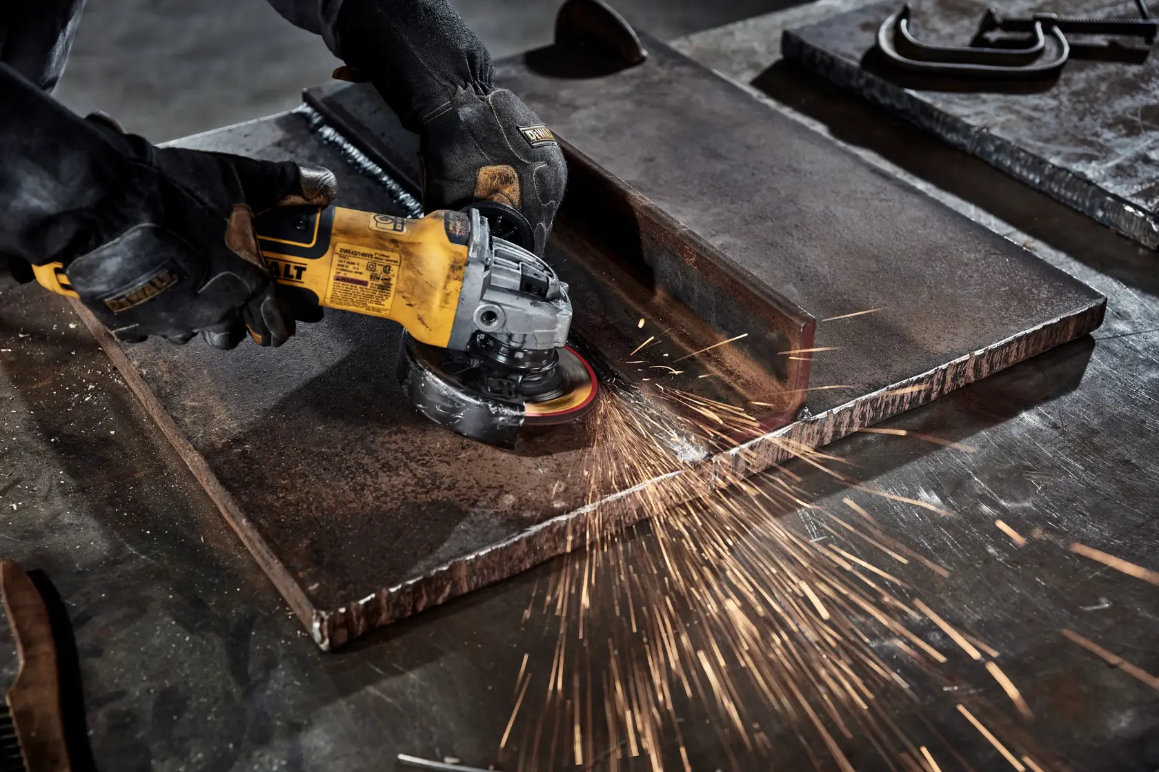 A person wearing protective gloves uses a DEWALT angle grinder to cut or grind a large metal sheet, producing sparks.
