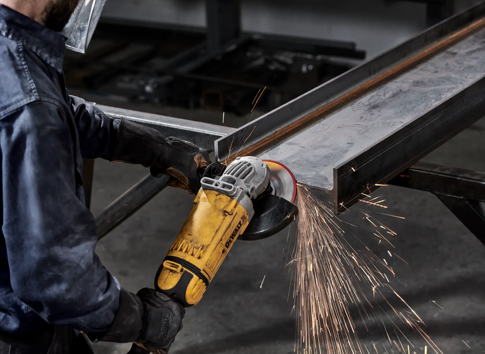 A person using a DEWALT angle grinder to grind or cut a piece of metal, producing sparks. The person wears protective gloves, a jacket, and a face shield for safety.