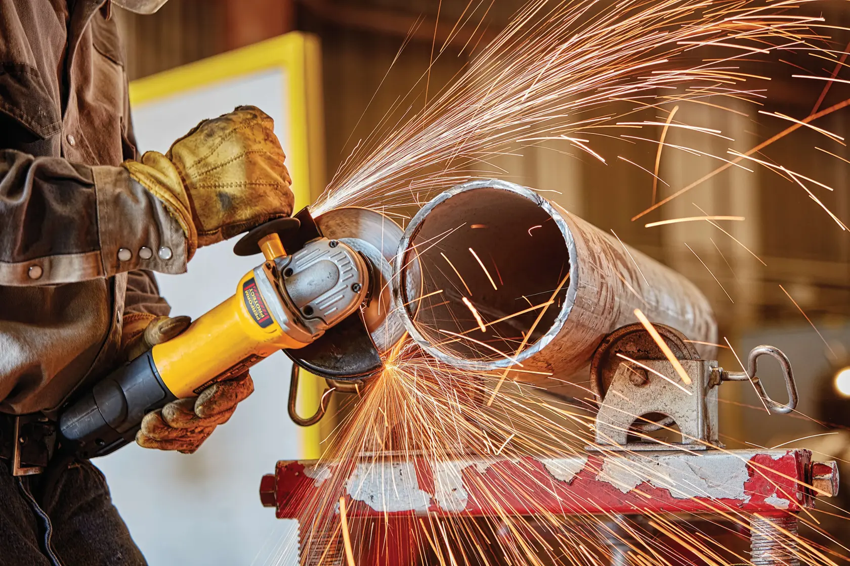 A person wearing gloves operates a DEWALT angle grinder, cutting a metal pipe and creating sparks.