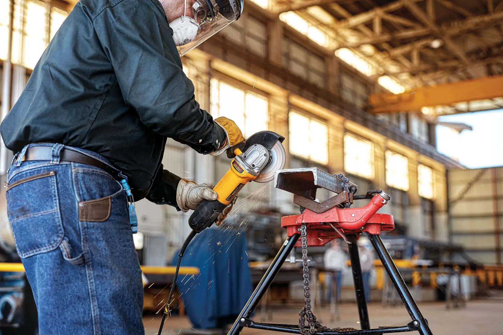 A person wearing safety gear uses a DEWALT angle grinder to cut metal on a workbench in an industrial workshop.