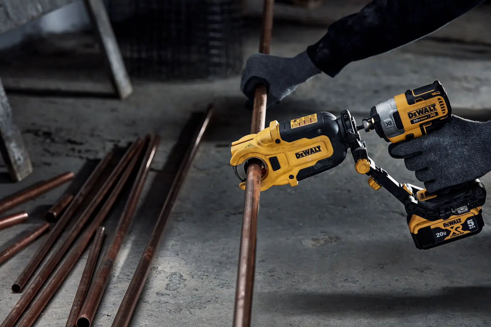 A person wearing gloves uses a DEWALT power tool to cut a copper pipe. Several copper pipes are lying on the ground nearby.