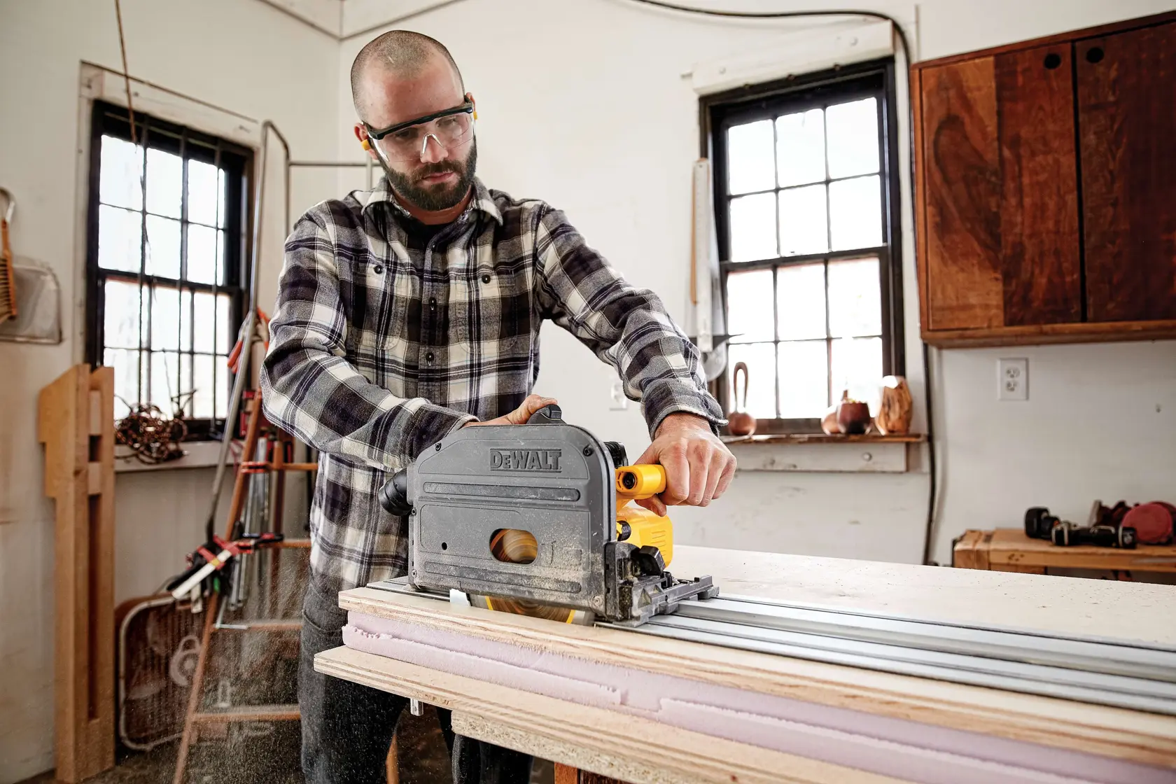 A person wearing a plaid shirt is using a DEWALT track saw to cut wood on a workbench in a workshop. The person's face is blurred for privacy.