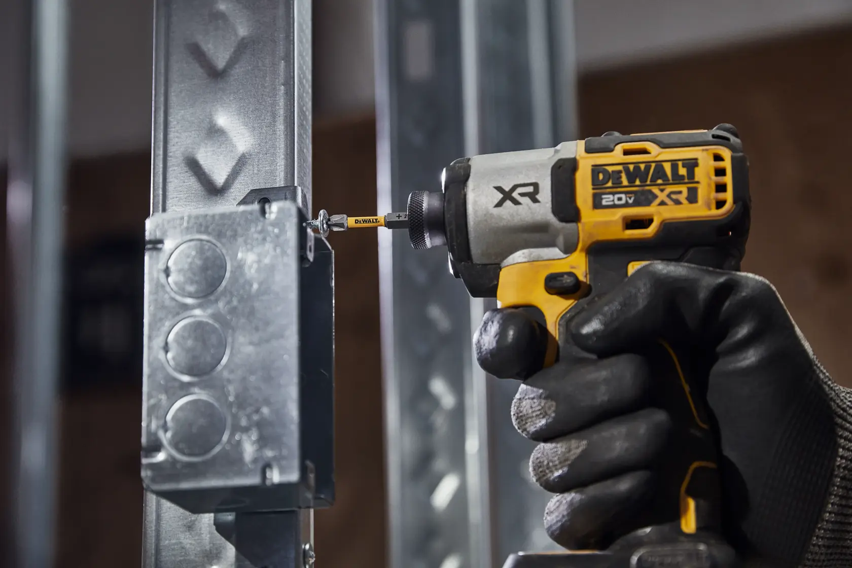 A gloved hand operates a DEWALT 20V XR cordless impact driver, driving a screw into a metal electrical box mounted on a steel frame.