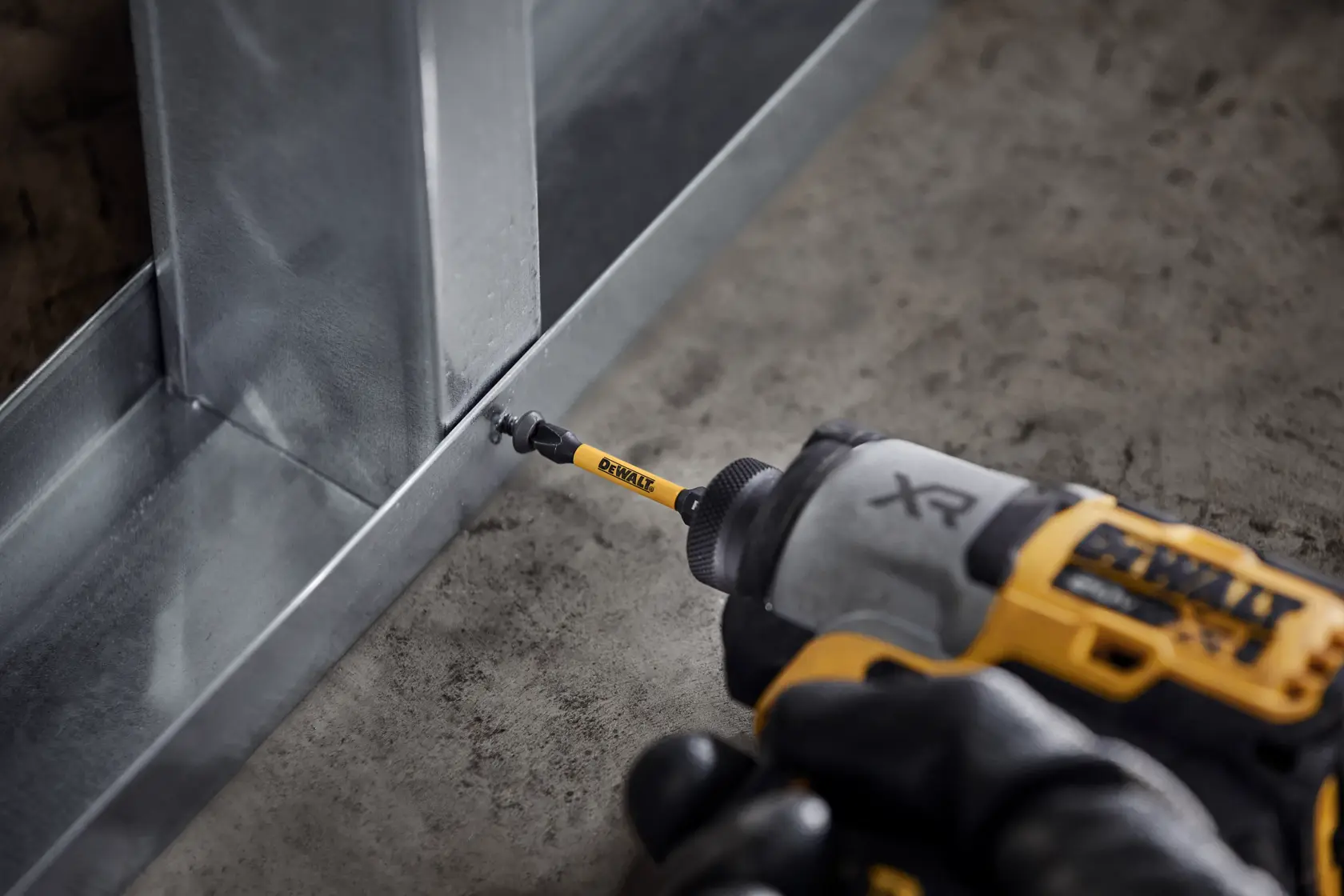 A close-up of a DEWALT cordless power drill with a screw driving bit installing a screw into a metal frame on a concrete floor.