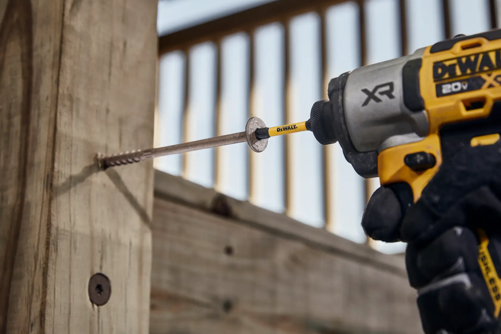 A DEWALT power drill is being used to drive a large screw into a piece of wood outdoors. The drill bit and screw are clearly visible.