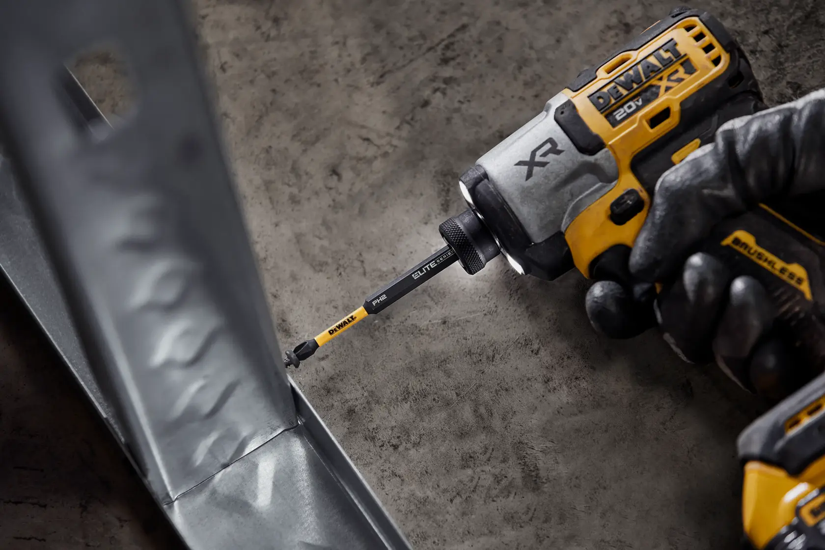 A close-up of a gloved hand using a DEWALT XR 20V cordless impact driver with a DEWALT Elite Series PH2 bit to fasten a screw into a metal bracket.