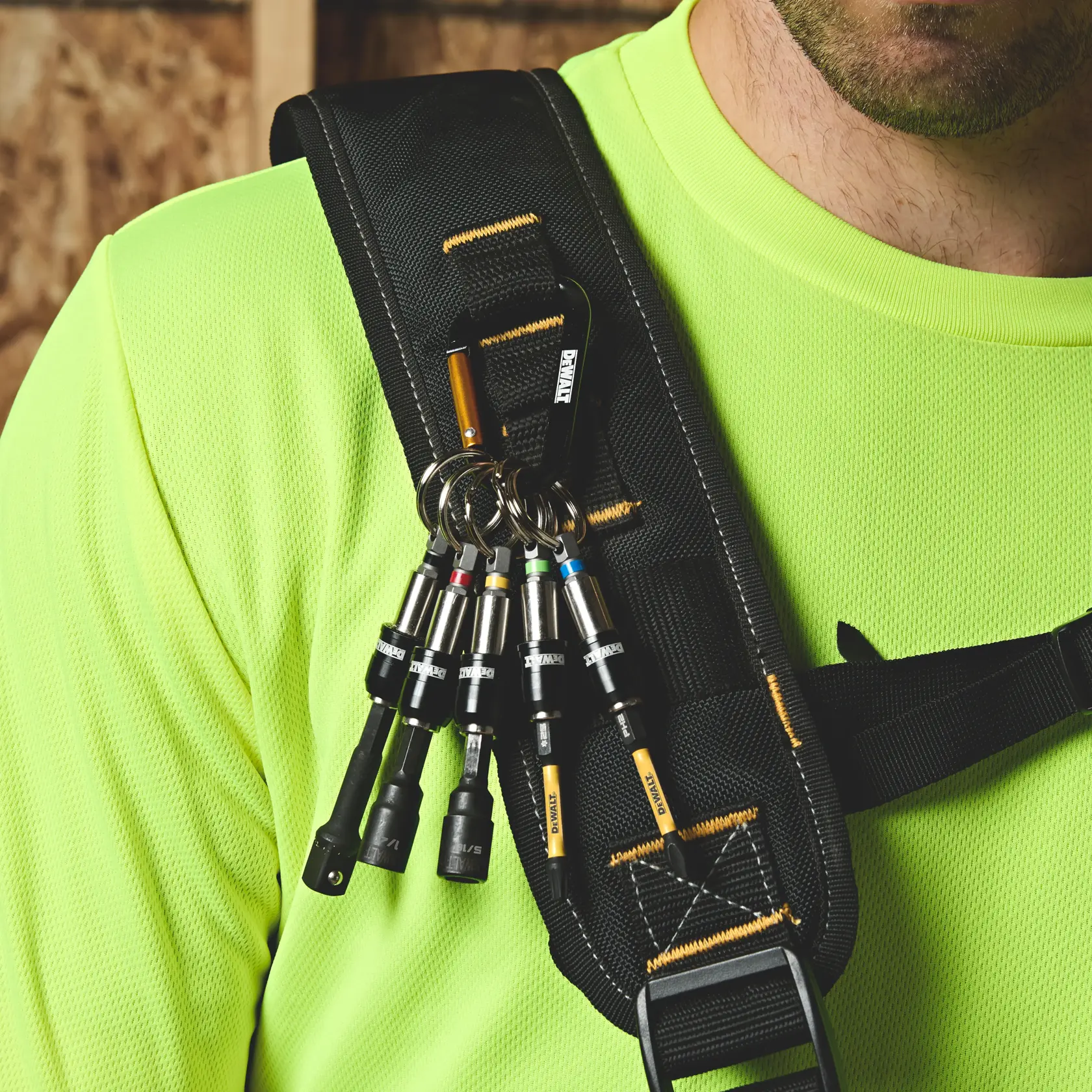 Close-up of a person wearing a neon yellow shirt and a black tool harness with a DEWALT branded carabiner attached to the shoulder strap, holding several DEWALT hex drill bit holders on key rings.