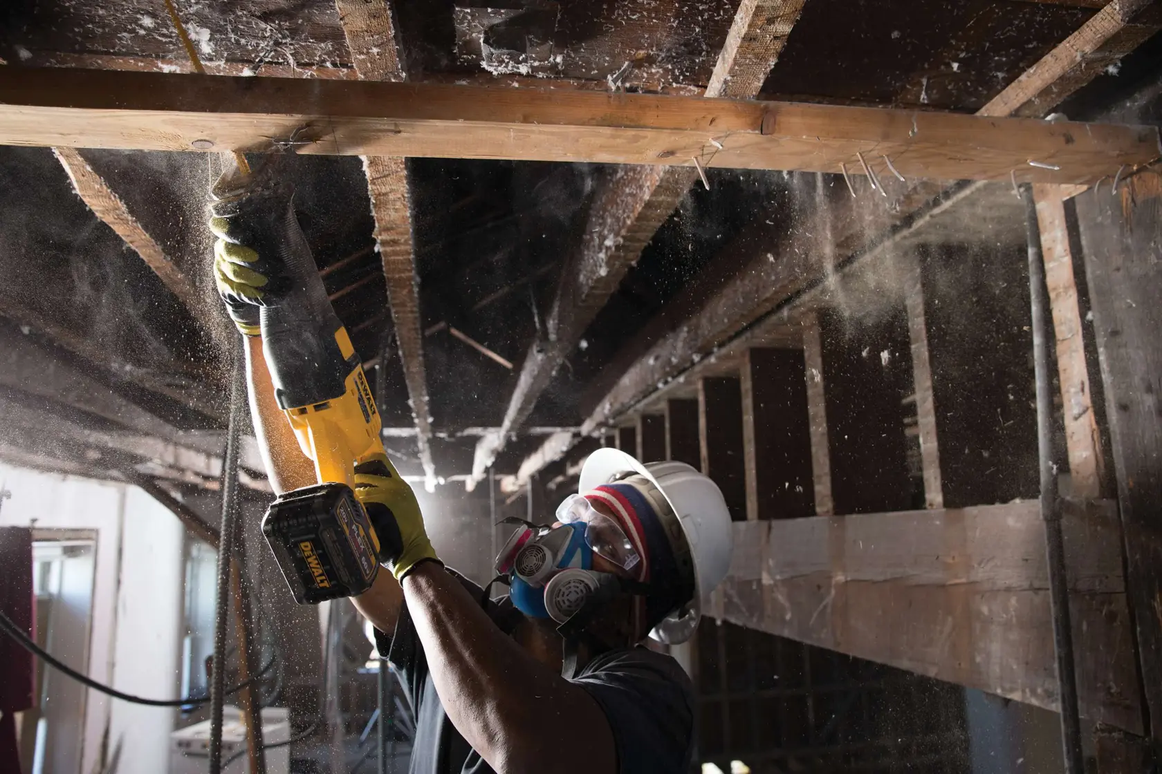A person wearing a hard hat, protective goggles, gloves, and a respirator mask is using a DEWALT cordless reciprocating saw to cut through a wooden beam in an industrial or construction setting. Dust particles are visible in the air.