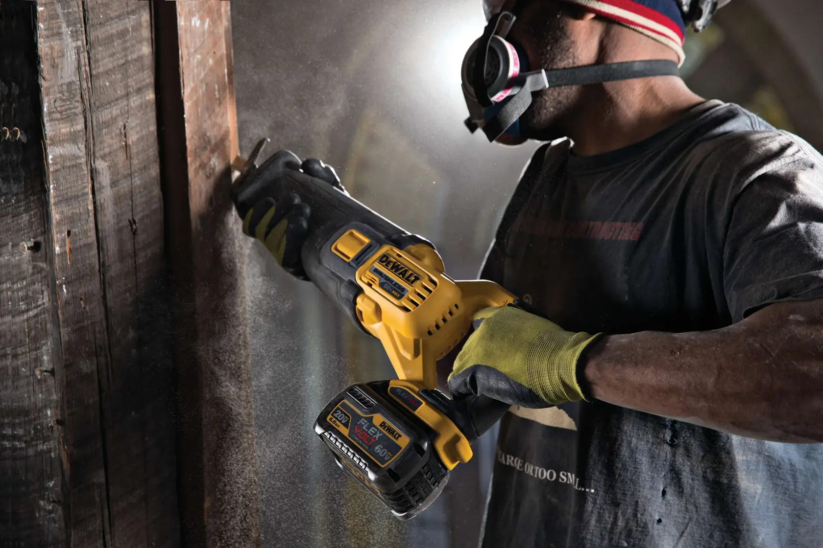 A person wearing safety gloves and a respirator mask is using a yellow and black DEWALT cordless reciprocating saw to cut through a wooden beam, with sawdust visible in the air.