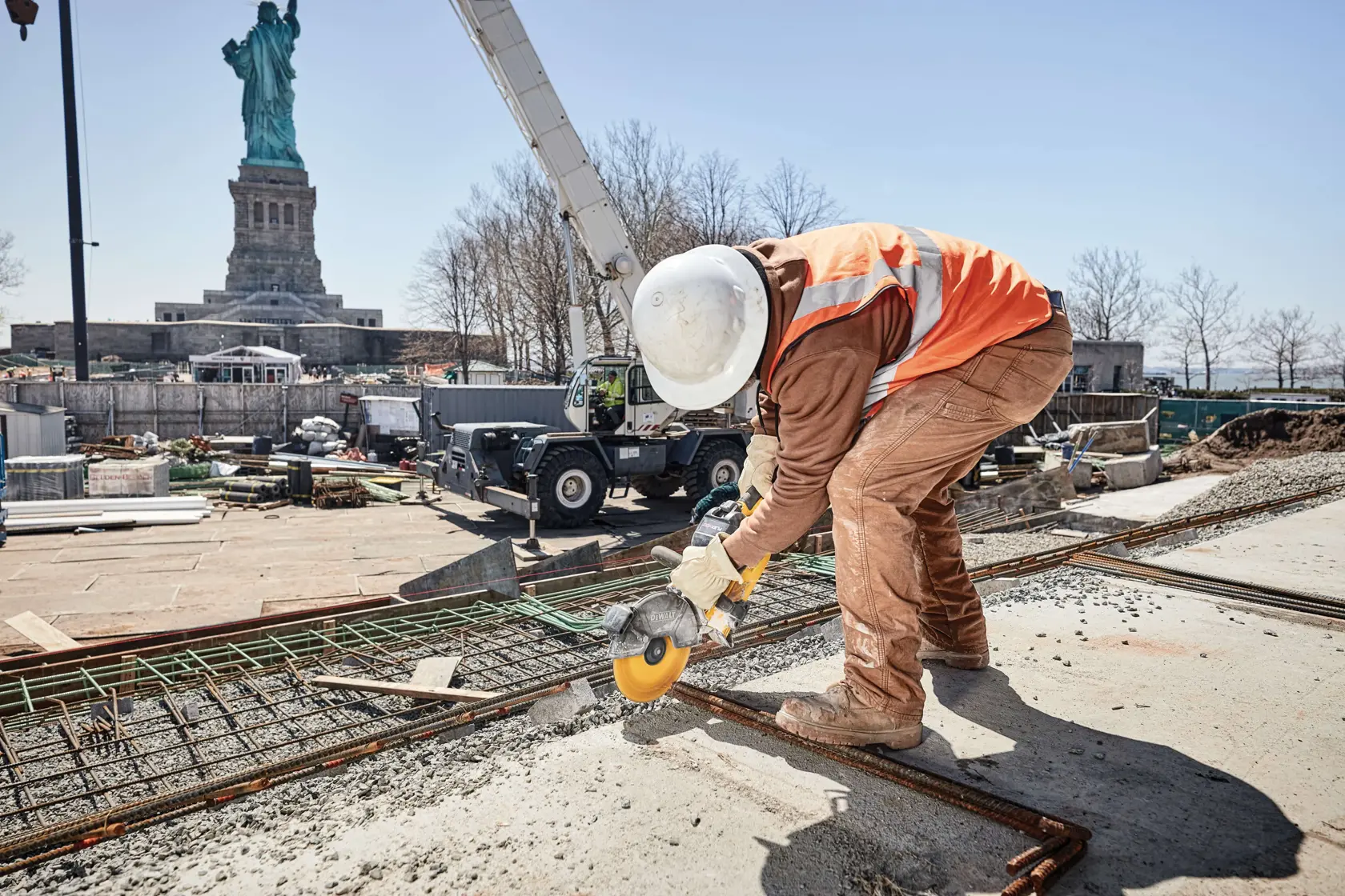 A construction worker wearing a safety helmet, gloves, and an orange reflective vest uses a yellow power tool to cut metal rebar on a construction site. The Statue of Liberty is visible in the background.