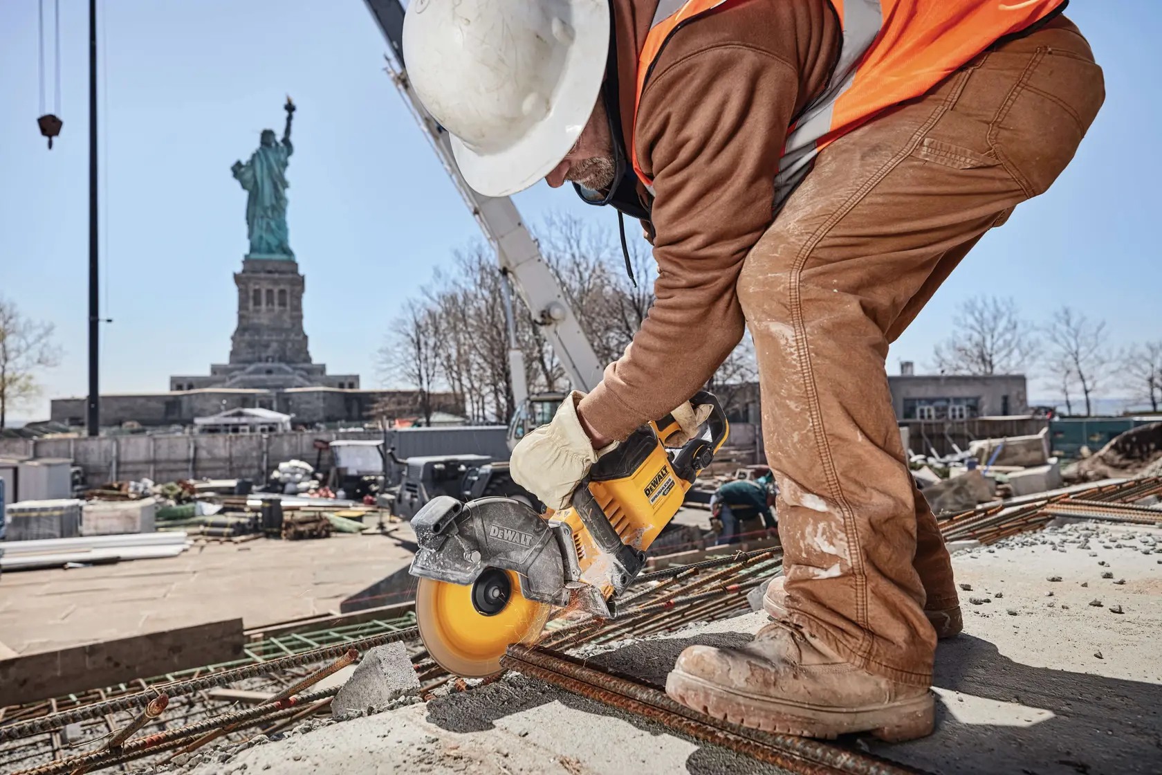 A person wearing a construction helmet and safety gear uses a yellow DEWALT power tool to cut through metal rods on a construction site, with the Statue of Liberty visible in the background.