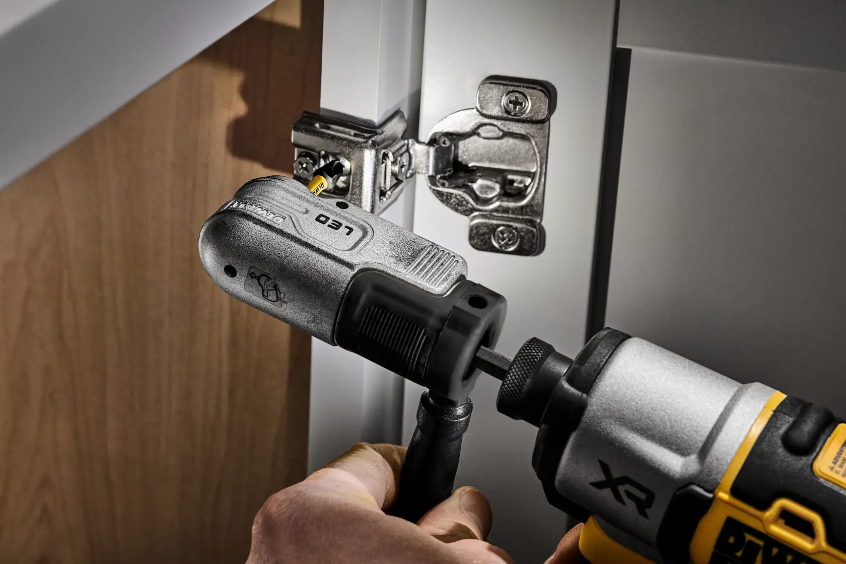 A DEWALT XR power drill with a right angle attachment is being used to install or tighten a screw on a cabinet hinge.