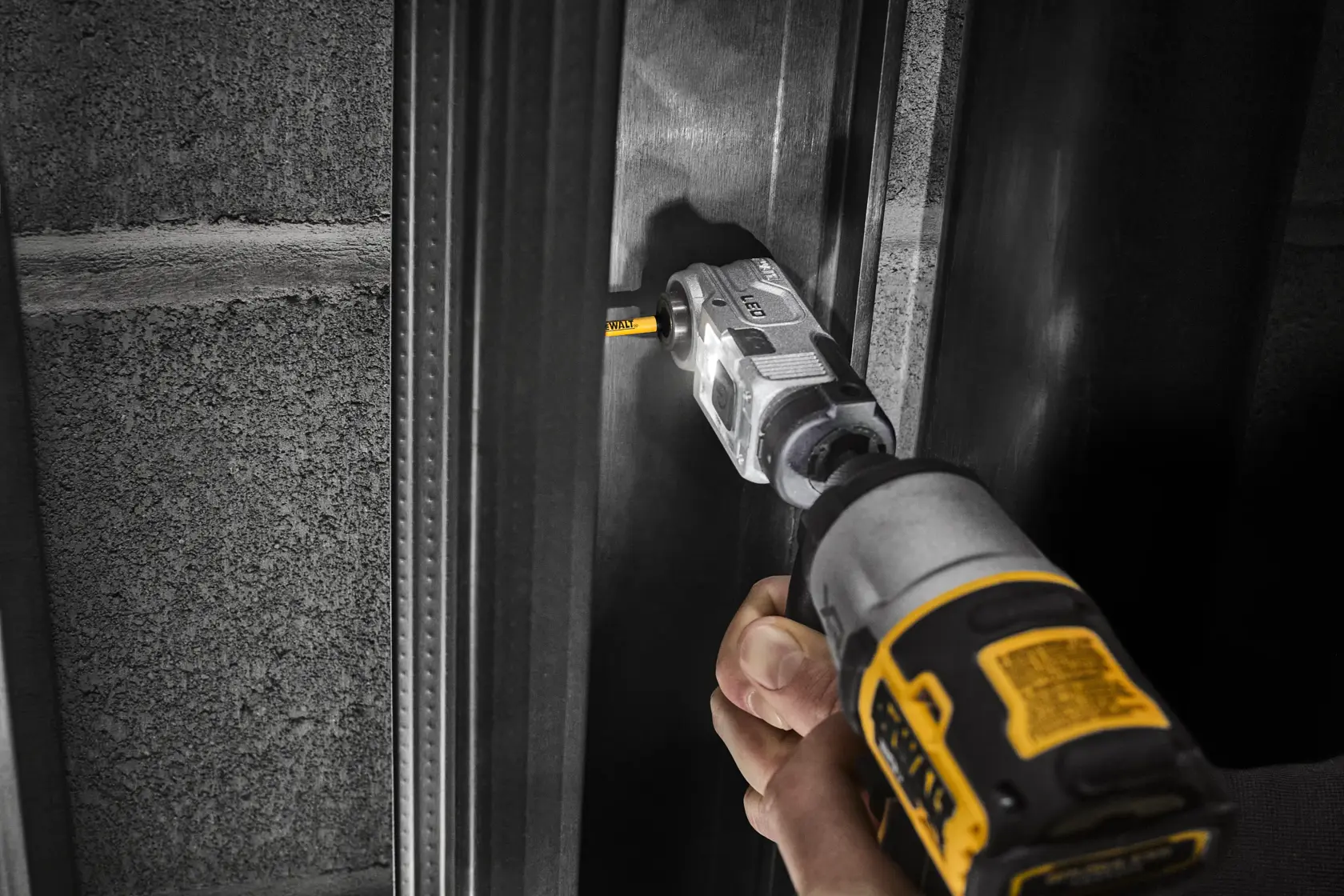 A hand using a DEWALT power tool with a right angle attachment to drill into a metal frame next to a concrete wall.