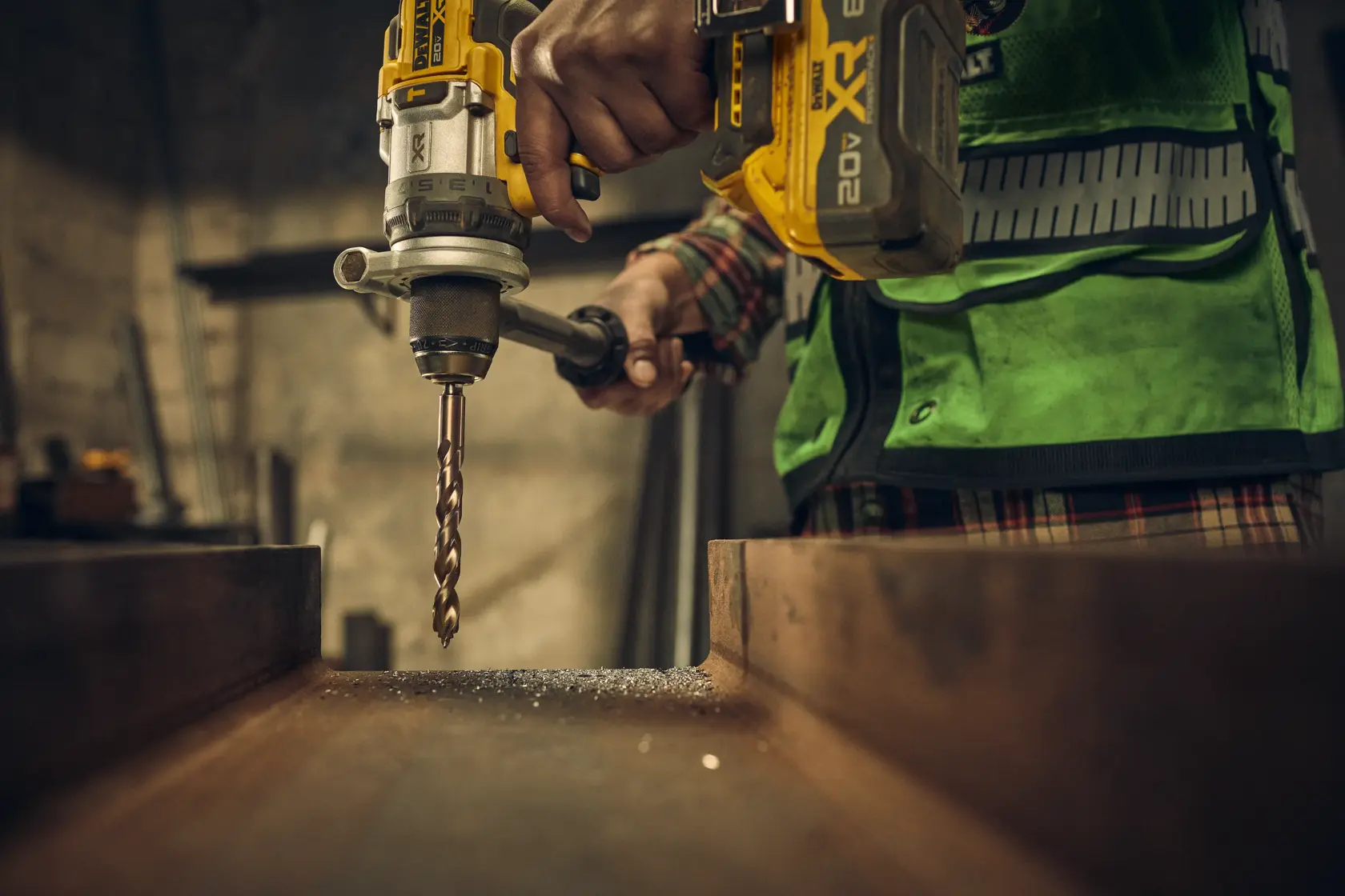 A person using a yellow DEWALT cordless drill with a metal drill bit to drill into a steel beam. The person is wearing a green safety vest and plaid shirt, and the scene appears to be in an industrial or workshop setting.