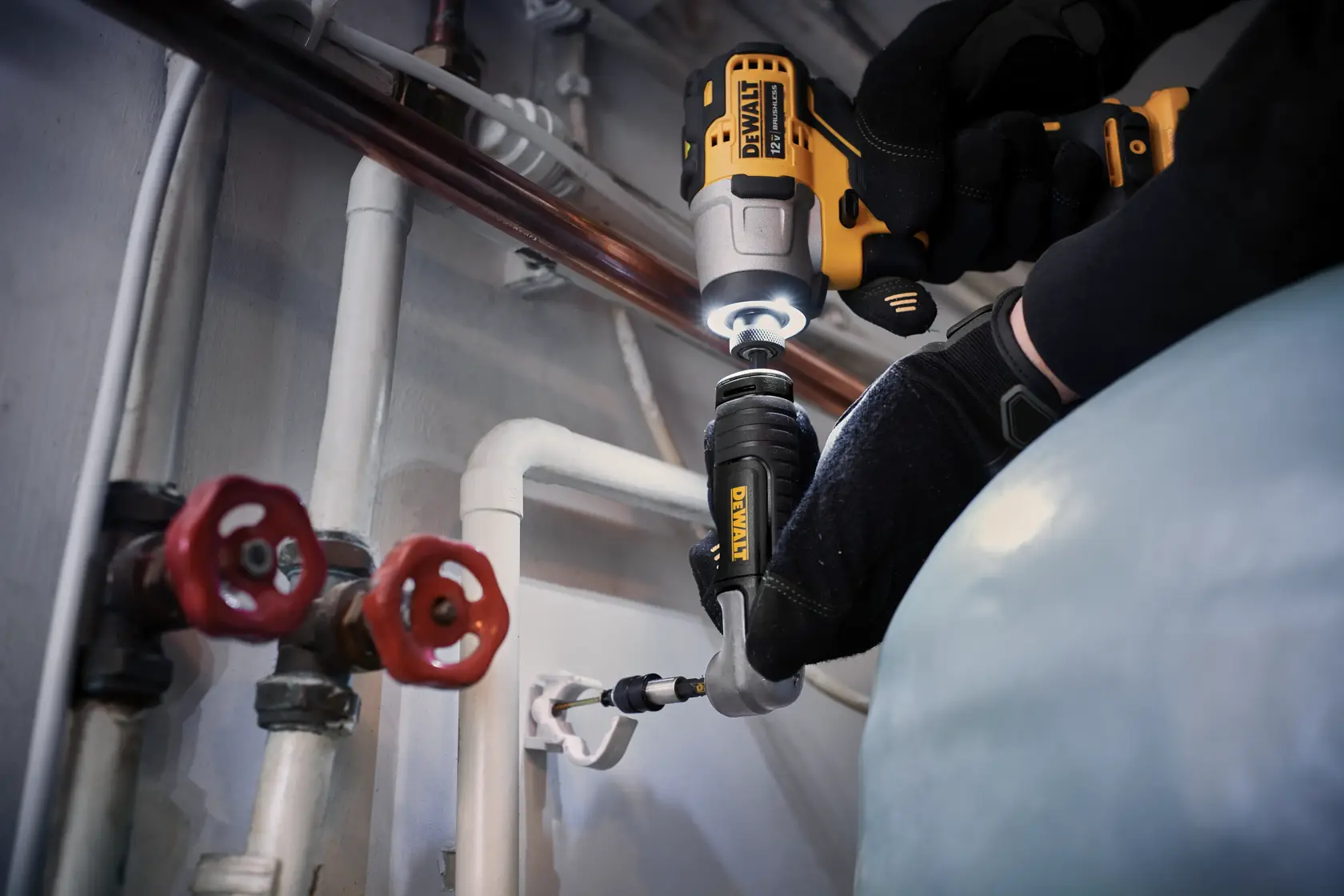 A person wearing black gloves uses a DEWALT cordless power drill with an attachment to work on piping in an industrial or utility setting. The scene includes visible pipes, valves, and fittings.