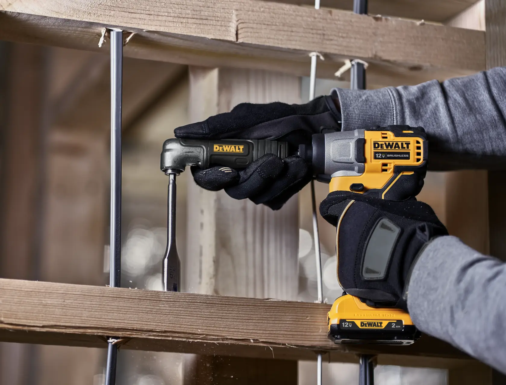 A person wearing black gloves uses a yellow DEWALT 12V cordless drill with a right angle attachment to drill into a wooden beam.