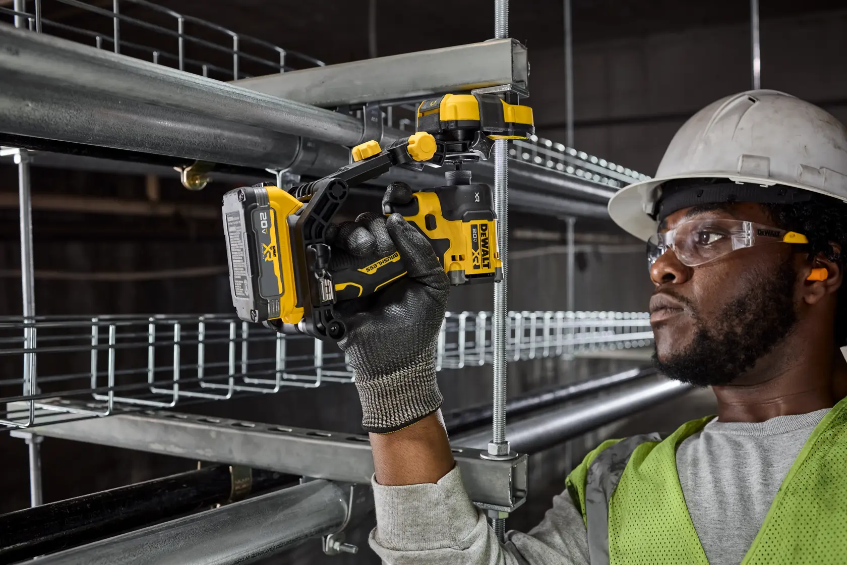 A person wearing safety gear and gloves uses a yellow and black DEWALT cordless power tool to fasten or drill into a metal pipe in a construction setting. The person's face is blurred for privacy.