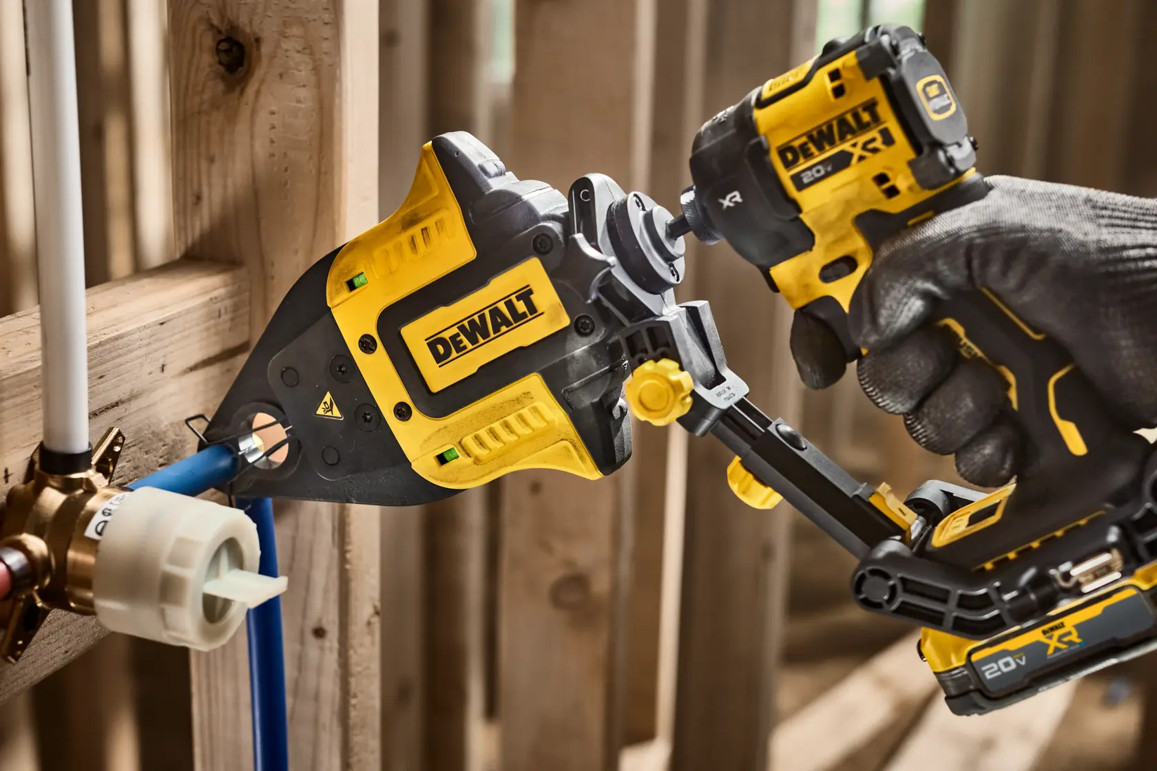 A DEWALT power tool is being used to press a pipe fitting onto a plumbing pipe, held by a gloved hand in a construction setting with wooden framing.