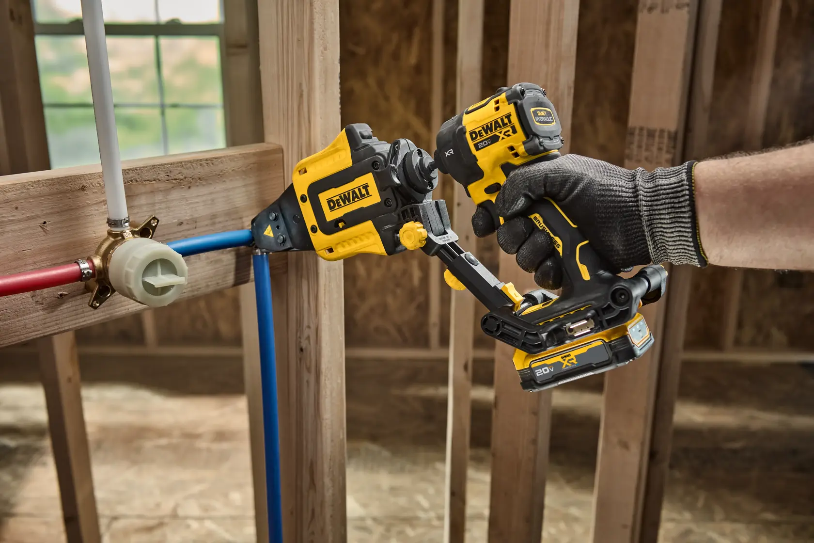 A gloved hand operates a DEWALT cordless power tool designed for pipe installation. The tool is being used on blue and red pipes inside a wooden framework of a building under construction.