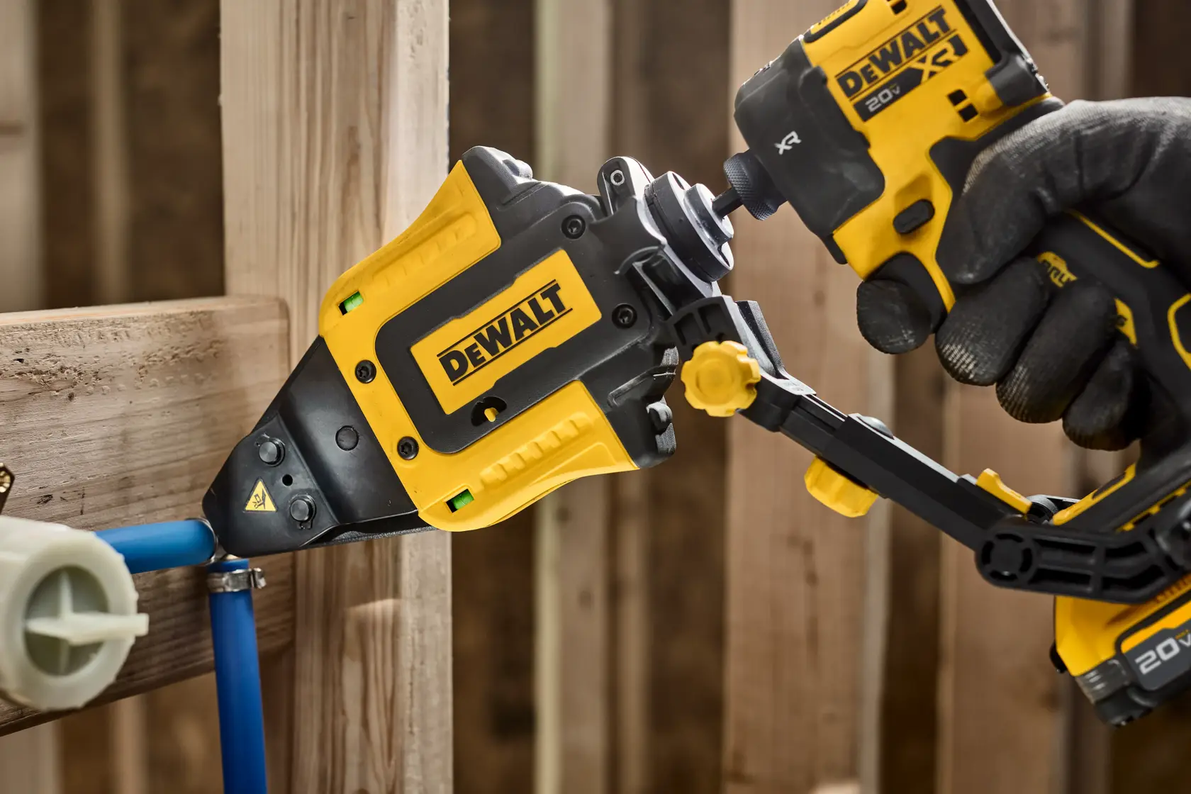 A close-up of a DEWALT power tool being used to fasten a pipe to a wooden frame. The tool is held by a gloved hand and is designed for plumbing or construction work.