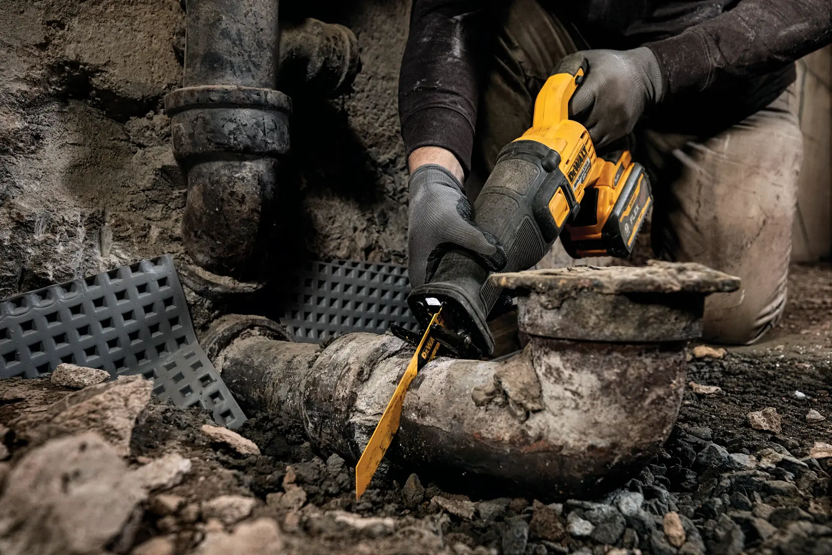 A person wearing gloves uses a DEWALT cordless reciprocating saw to cut a large, rusty metal pipe in an industrial or construction setting.