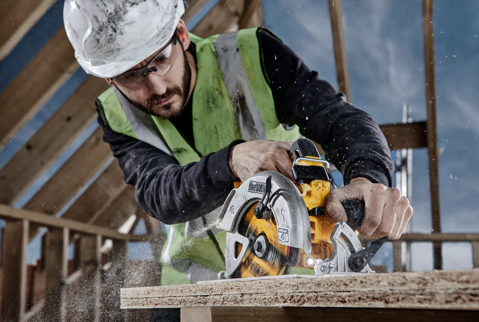 A construction worker wearing a safety vest and helmet uses a DEWALT circular saw to cut through a wooden board at a building site, with sawdust flying around.