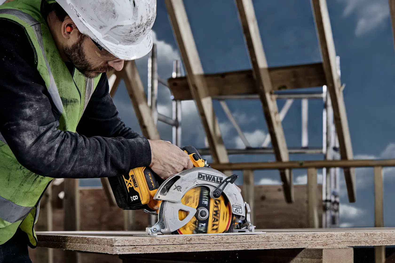 A person wearing a safety vest and helmet is using a DEWALT cordless circular saw to cut a wooden board at an outdoor construction site.