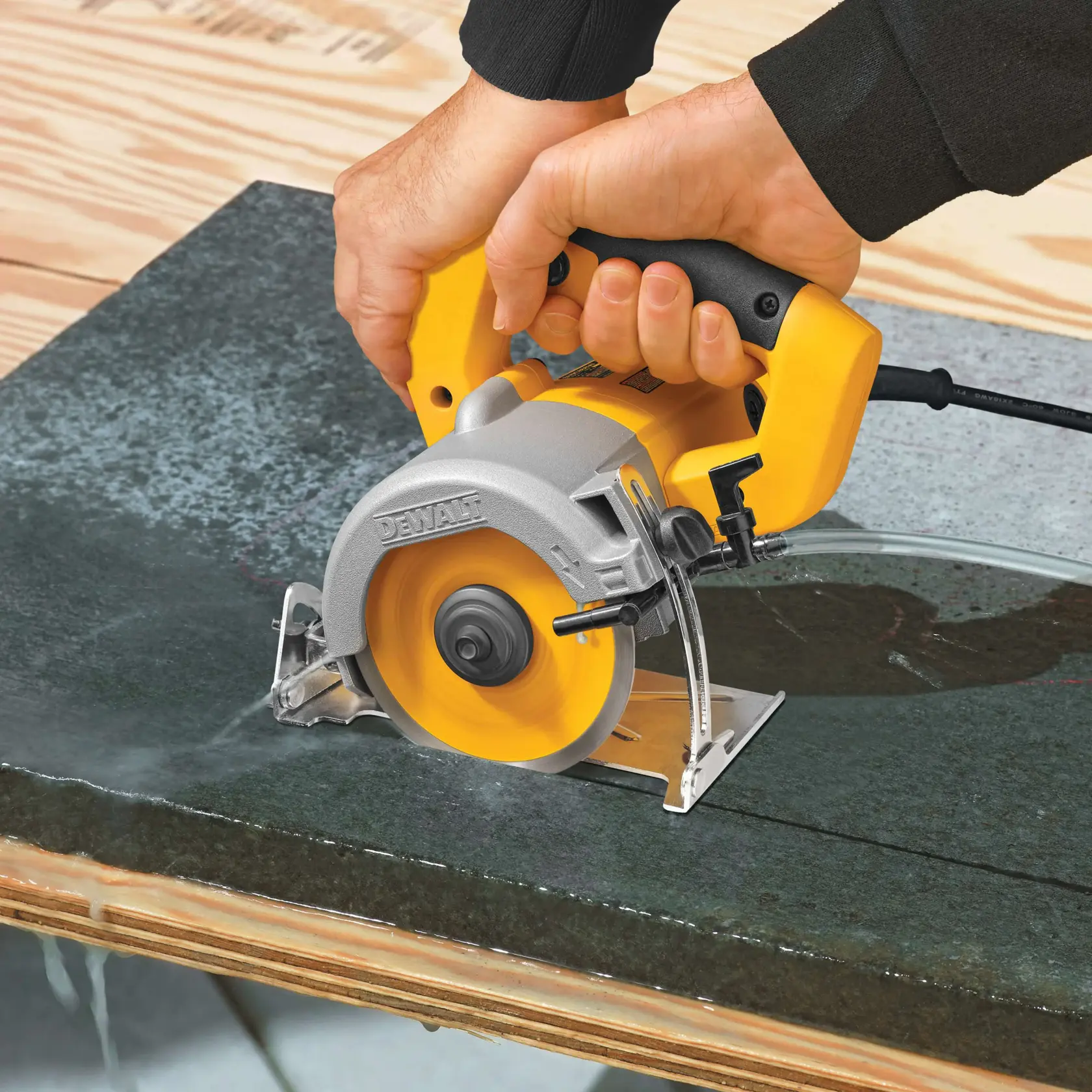 A person using a DEWALT wet tile saw to cut a piece of dark stone or tile on a wooden work surface. Water is being used to cool the blade.
