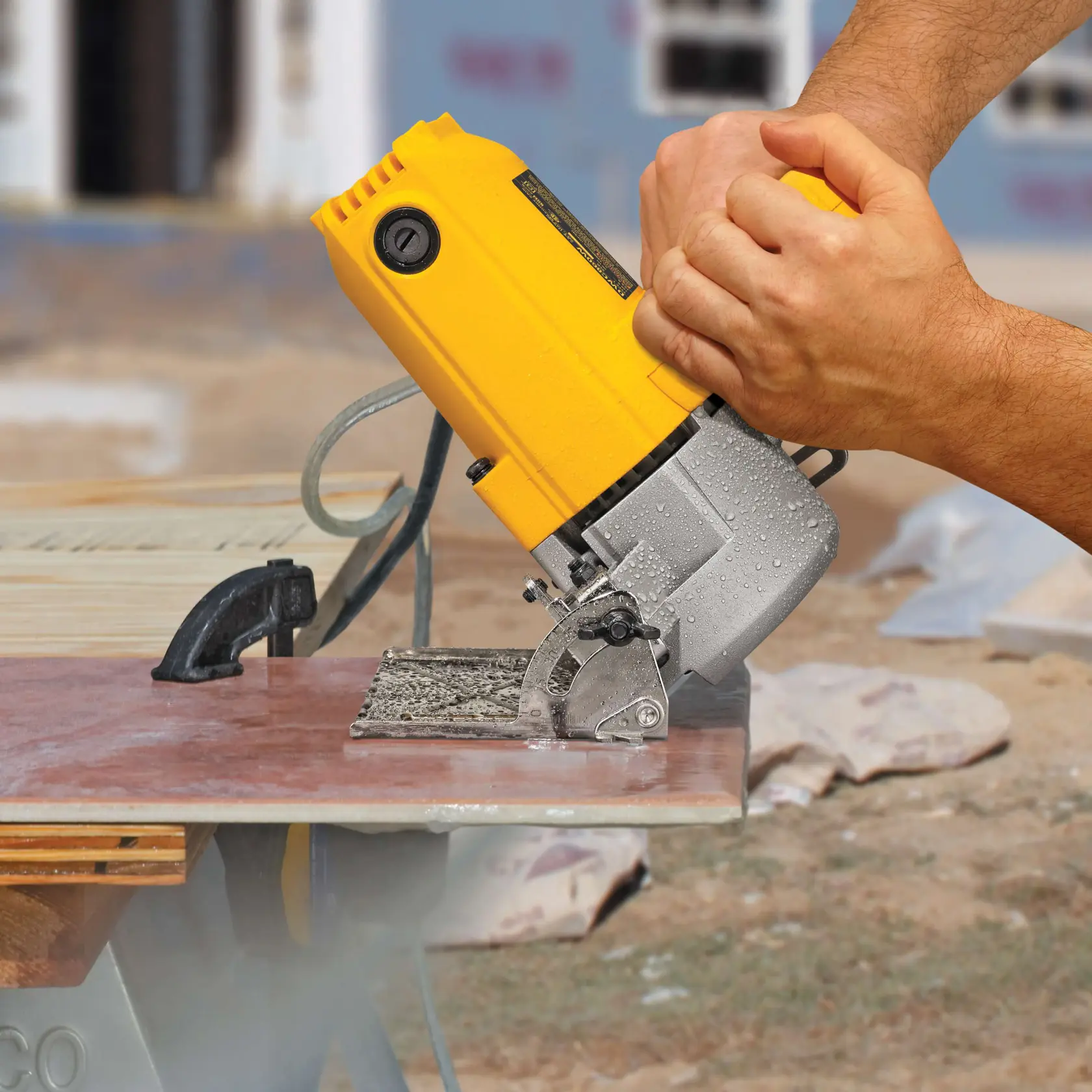 A close-up of hands operating a yellow DEWALT tile cutter, cutting a ceramic tile on a workbench outdoors.