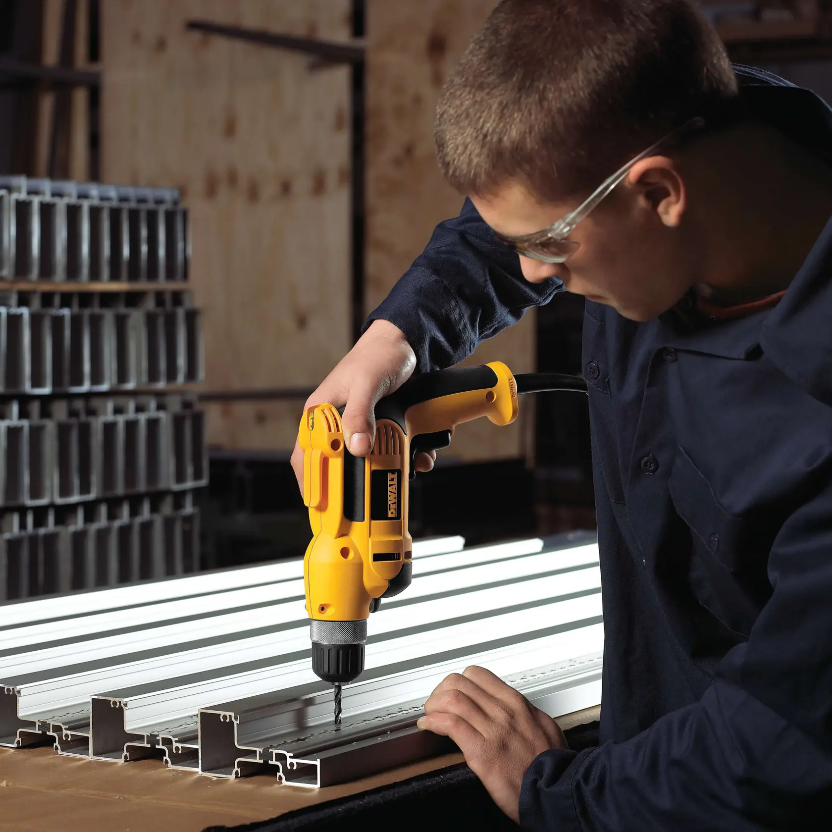 A person using a yellow DEWALT electric drill to drill into metal profiles placed on a workbench. The person is wearing a dark work shirt and safety glasses. The background includes stacks of metal sections and a workshop setting.