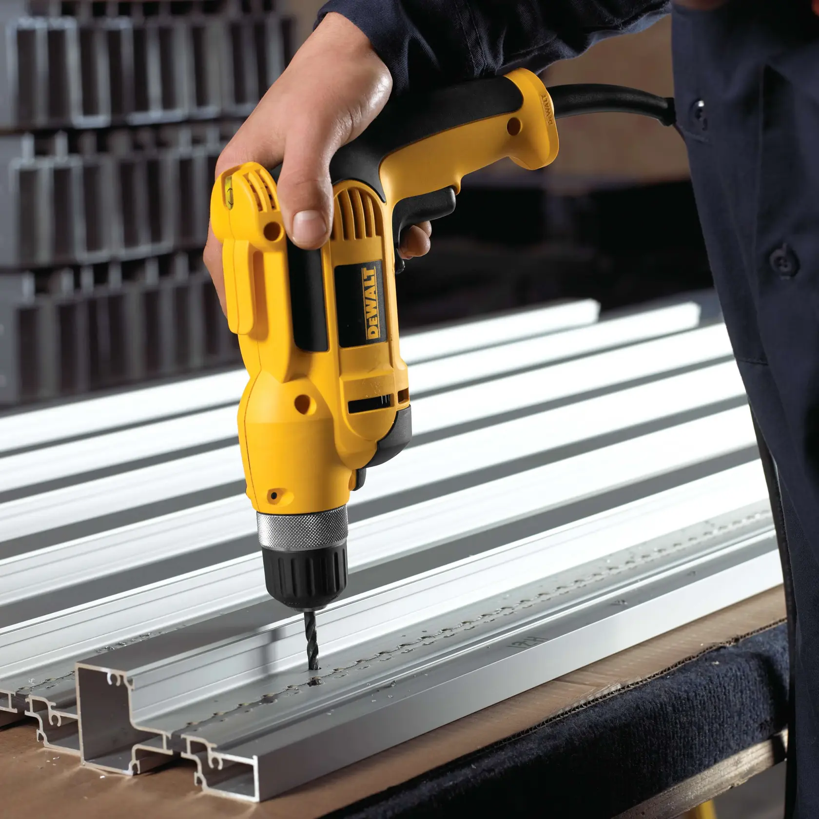 A person is using a yellow DEWALT electric drill to make holes in aluminum metal channels that are laid out on a workbench.