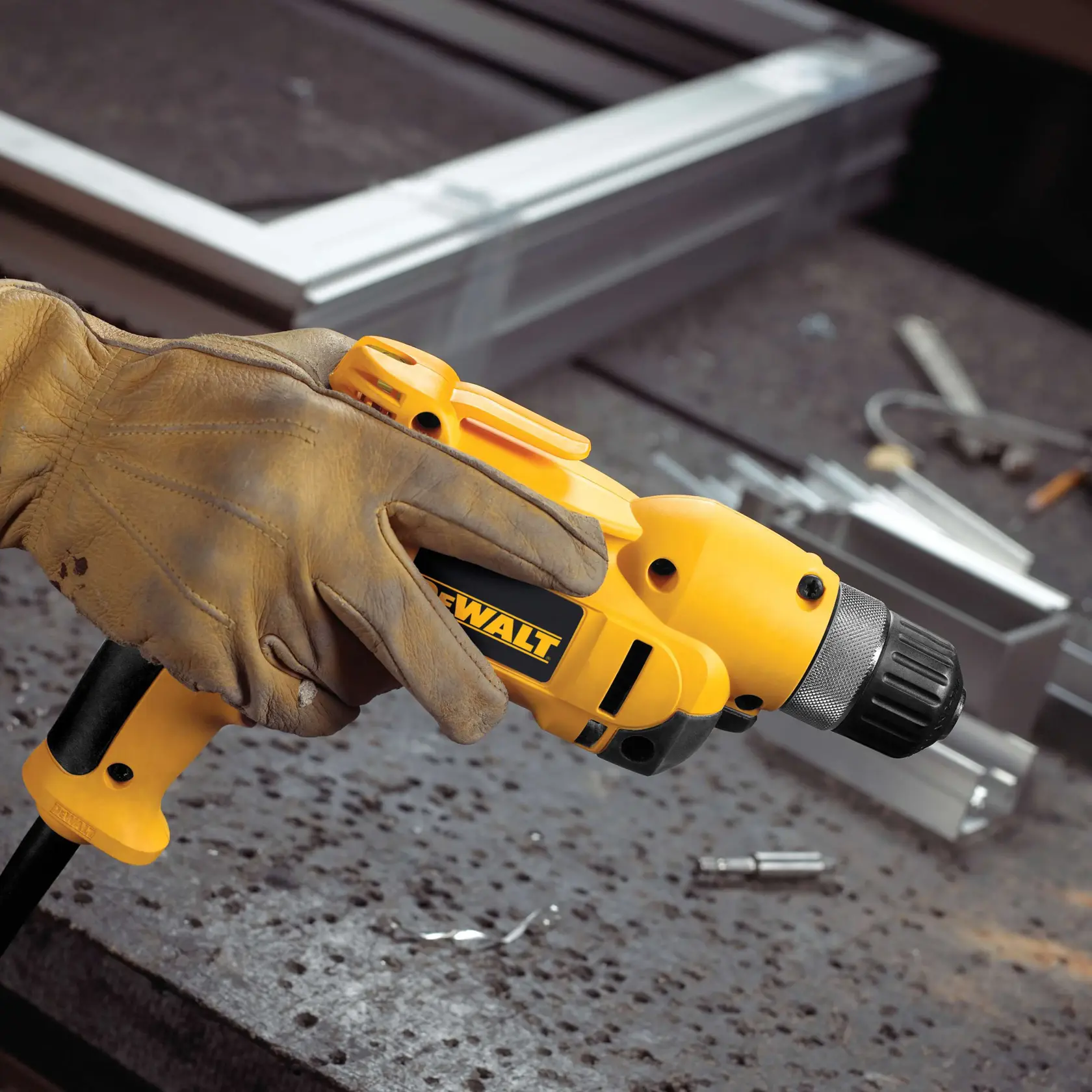 A gloved hand holding a yellow DEWALT corded drill, positioned above a workbench with metal parts and tools in the background.